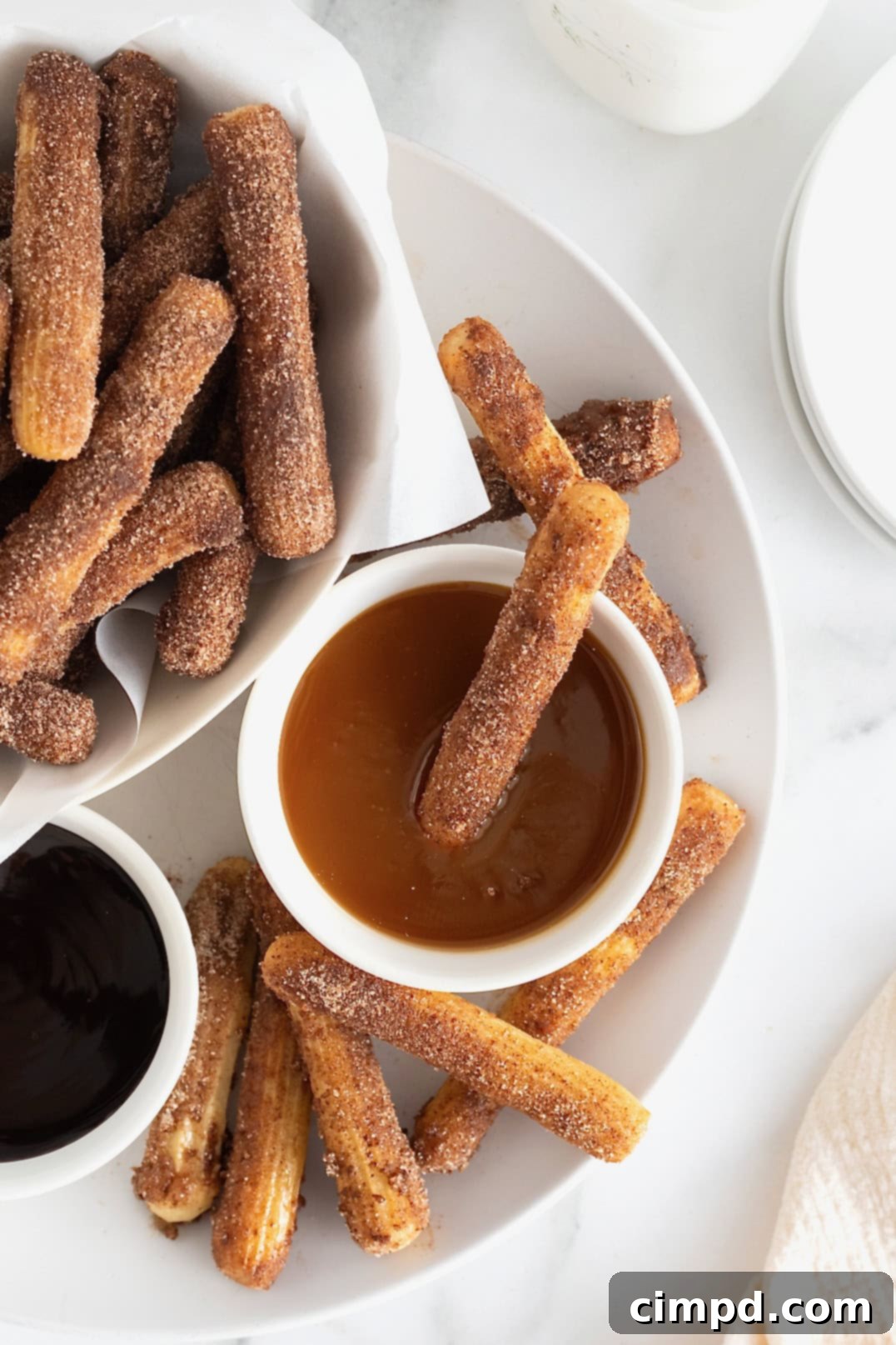 Guilt-Free Baked Churros 5 A parchment lined bowl of baked churros next to a small white dish of chocolate sauce and a small white dish of caramel sauce on a large white platter. A single churro is sticking out of the caramel sauce.