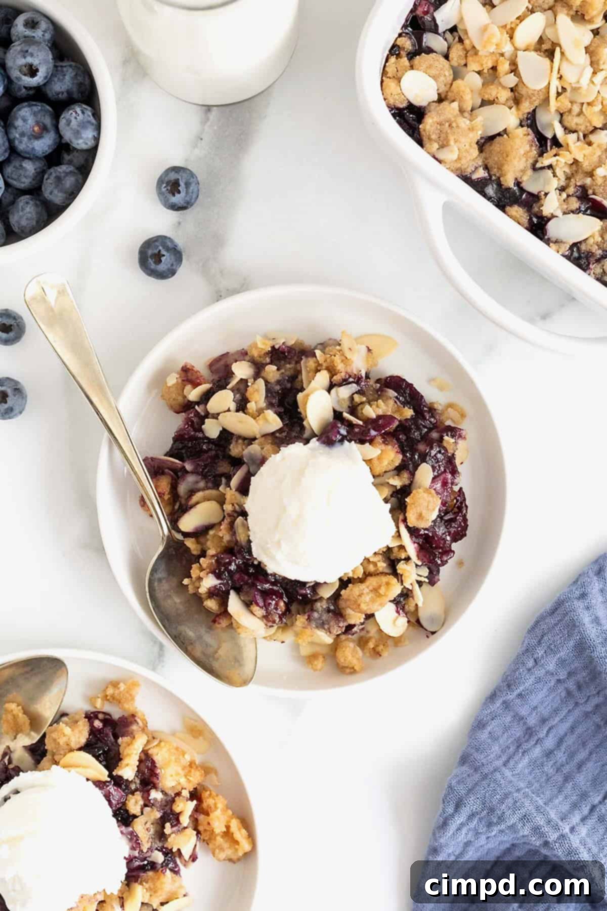 A small white plate of blueberry almond crisp. There is a scoop of vanilla ice-cream on the crisp and a spoon lying next to it.