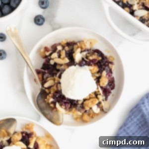 A small white plate of blueberry almond crisp. There is a scoop of vanilla ice-cream on the crisp and a spoon lying next to it.