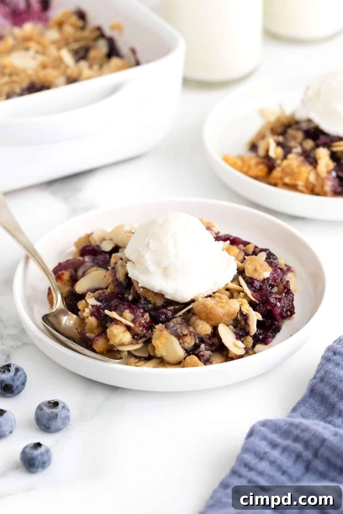 Two small white plates of blueberry almond crisp. There is a scoop of vanilla ice-cream on the crisp and a spoon lying next to it.