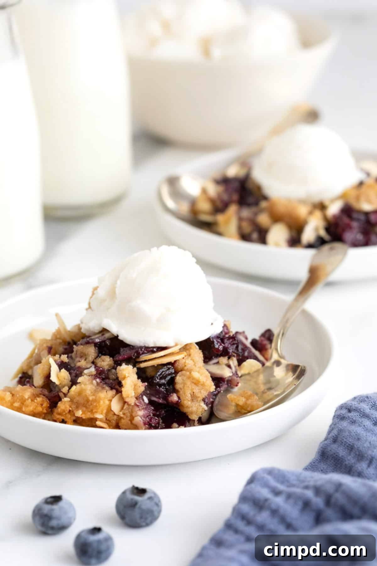 Two small white plates of blueberry almond crisp. There is a scoop of vanilla ice-cream on the crisp and a spoon lying next to it.