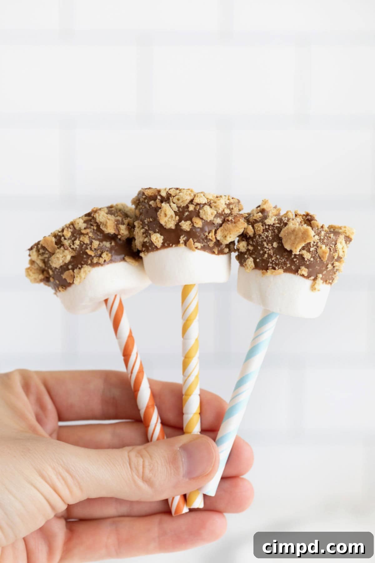 A hand holding three s'mores marshmallow pops on pastel and white striped straws.