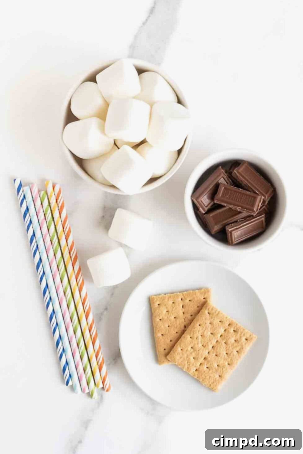 Ingredients to make s'mores marshmallow pops in white dishes on a white marble counter.