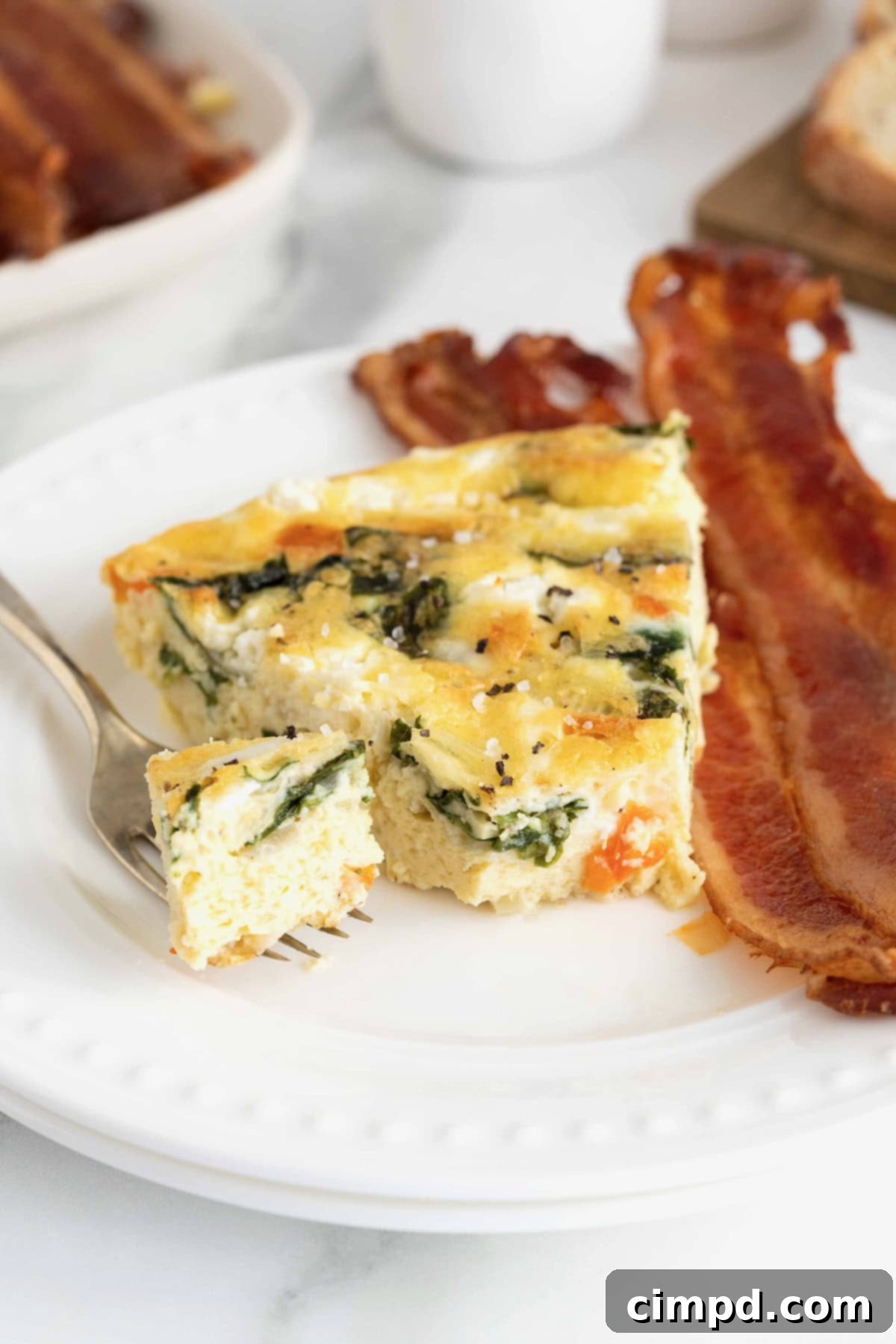 A perfectly portioned slice of frittata on a white plate, accompanied by three strips of crispy bacon, highlighting its fluffy texture.