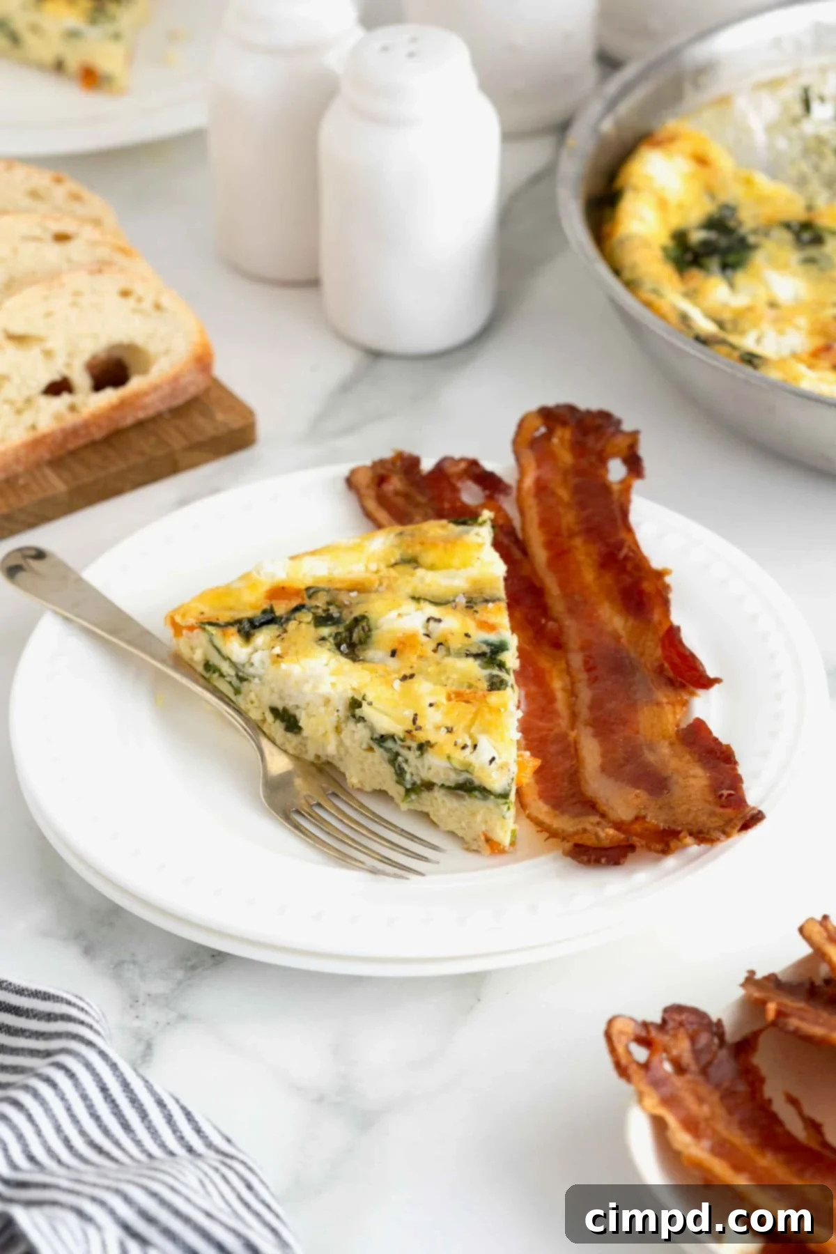 A close-up of a frittata slice on a white plate, alongside two pieces of bacon, showcasing the vibrant red peppers and green spinach.