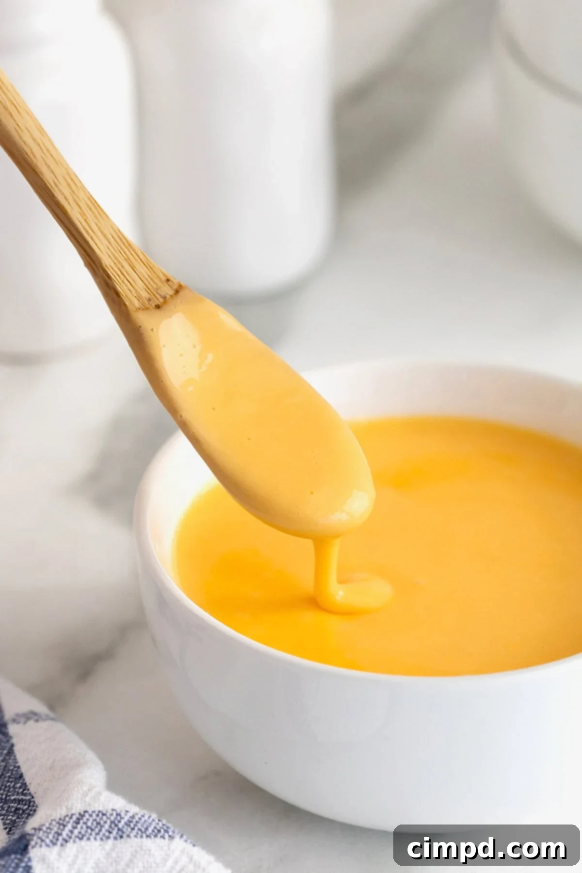 A wooden spoonful of golden cheese sauce dripping lusciously into a white bowl of more cheese sauce, highlighting its thick, smooth consistency.