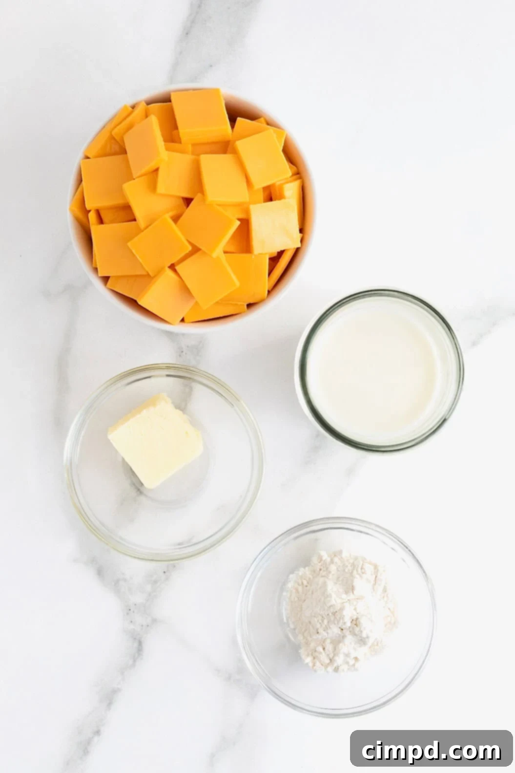 Neatly arranged ingredients for easy cheese sauce in small clear glass dishes on a pristine white marble counter, ready for preparation.