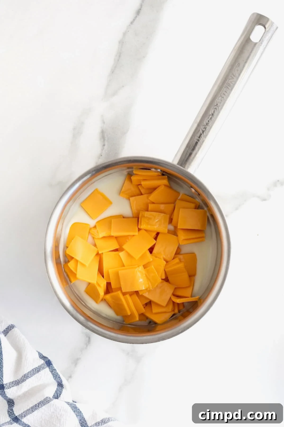 Squares of American cheese being added to a metal saucepan filled with the thickening roux and milk mixture.