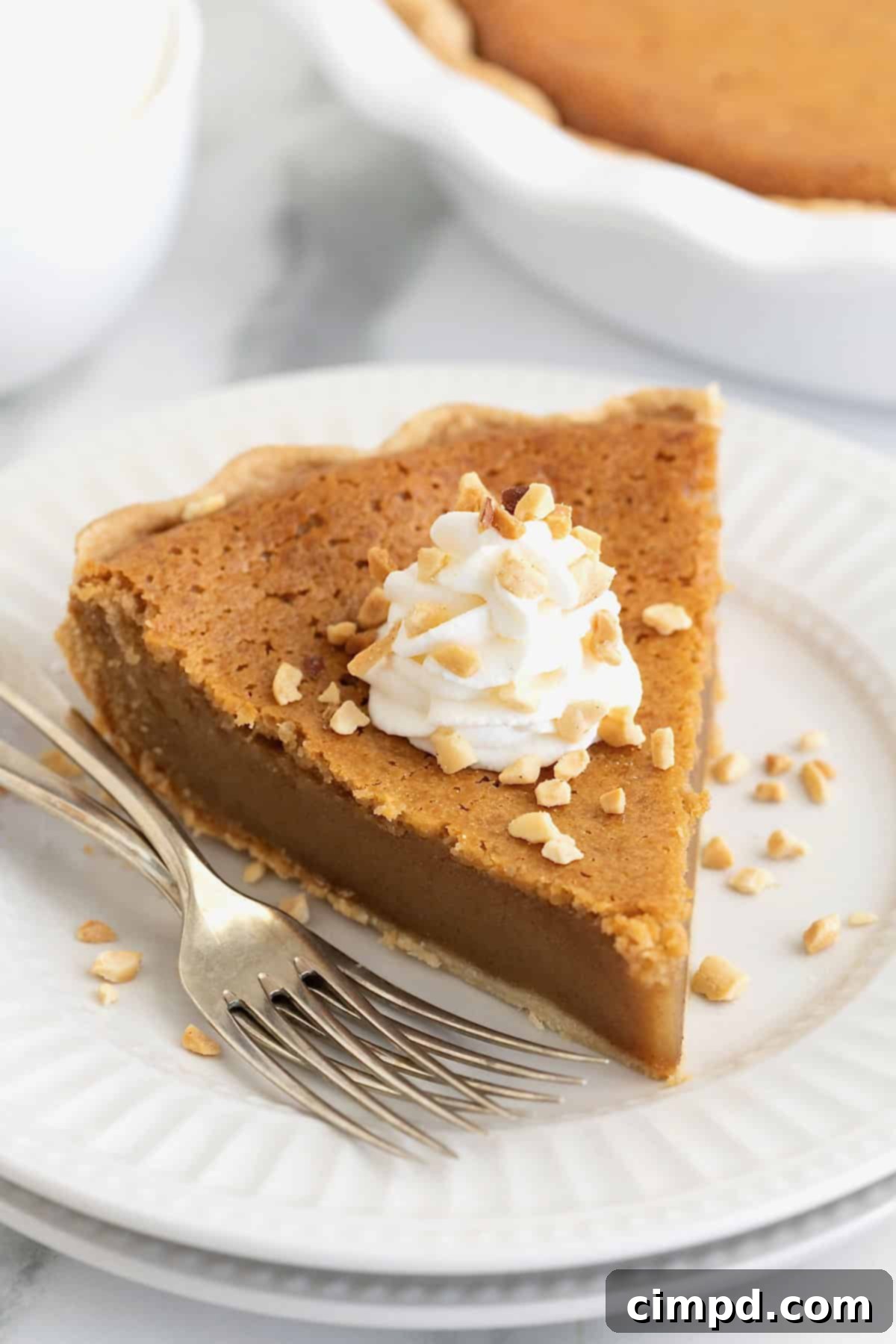 A generous slice of Baked Peanut Butter Pie, elegantly topped with a dollop of fluffy whipped cream and a sprinkle of chopped peanuts, rests on a pristine white plate, accompanied by two forks.