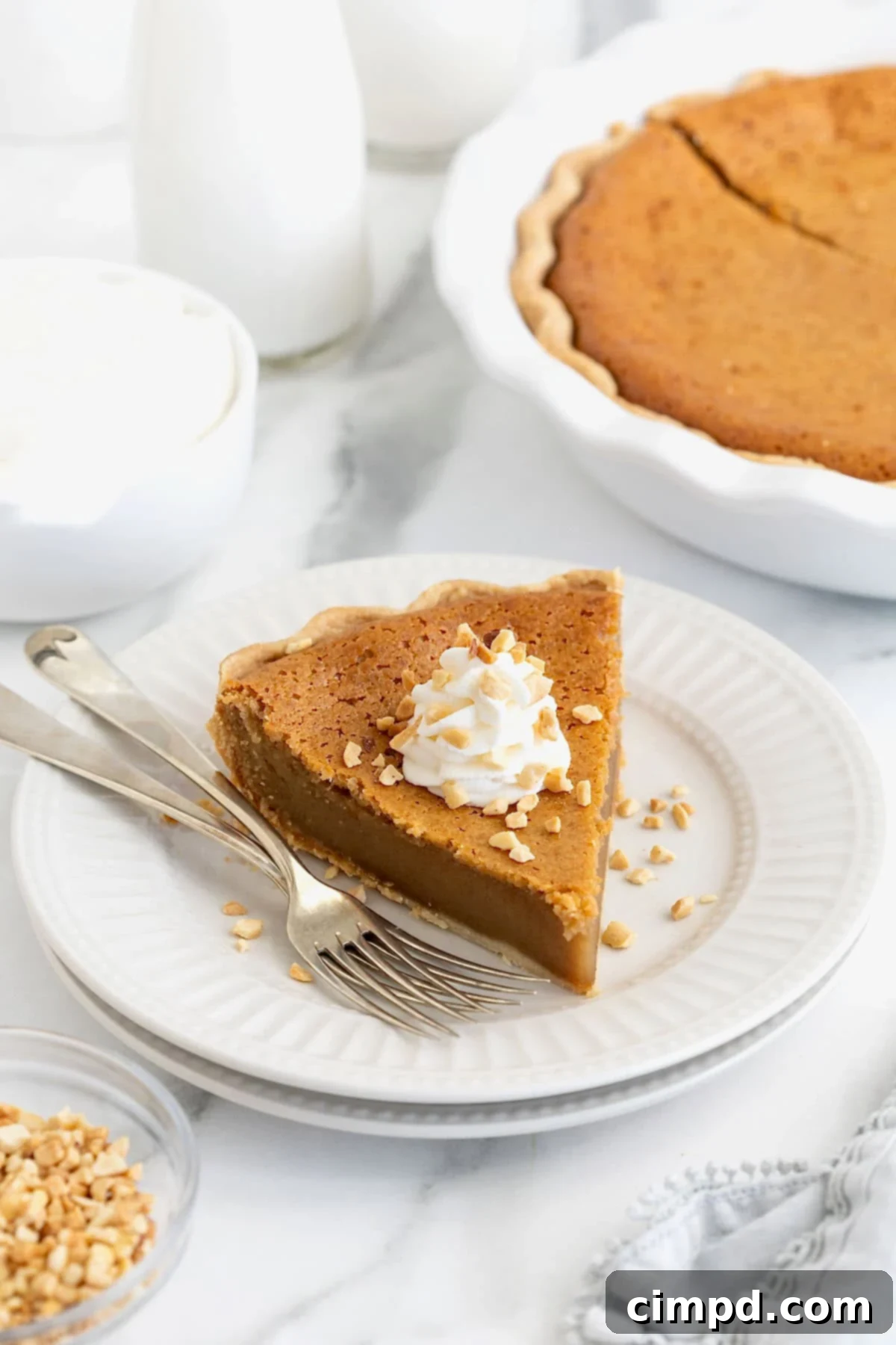 A ready-to-serve slice of peanut butter pie, adorned with whipped cream and chopped peanuts, presented on a white plate with two forks beside it.
