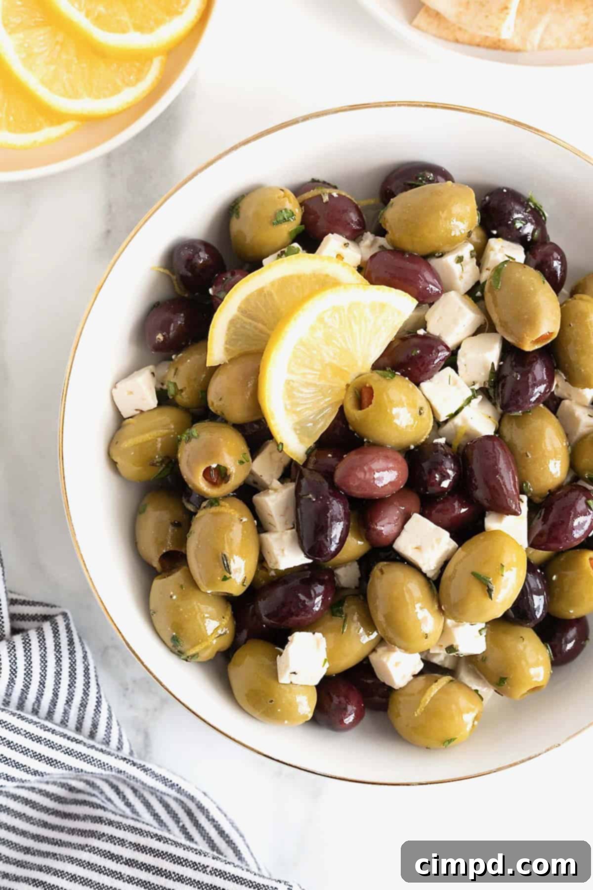 A gold rimmed white serving bowl filled with green and purple olives and small cubes of feta, ready to be served.