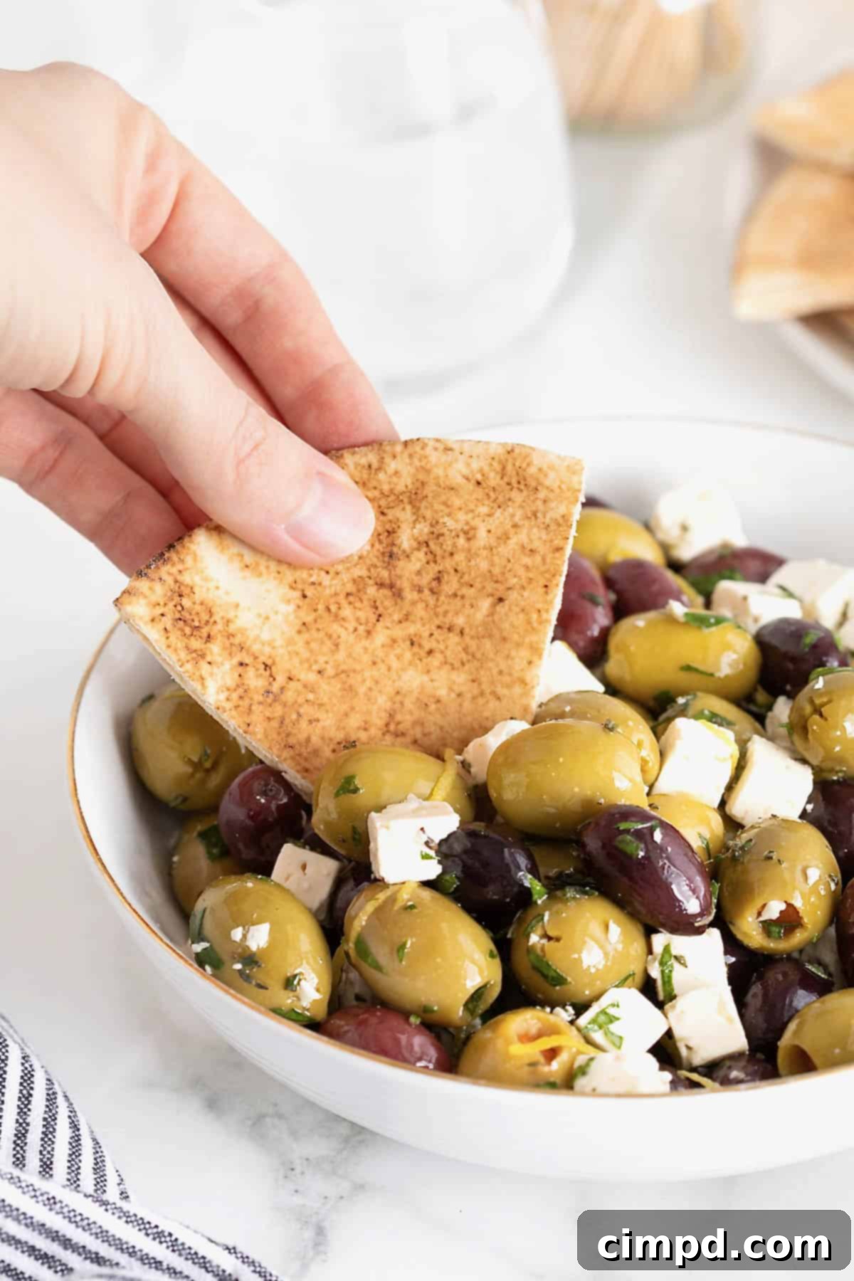 Marinated olives and feta being scooped onto a pita chip from a gold-rimmed white bowl, highlighting the serving suggestion.