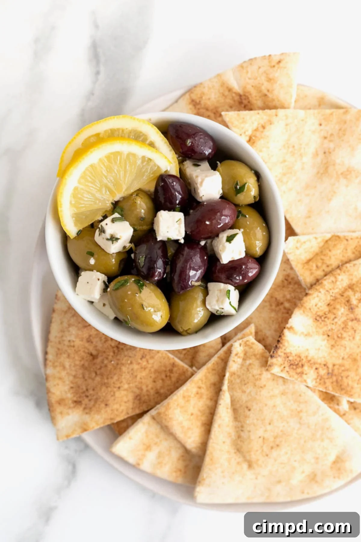 A bowl of marinated olives and feta surrounded by golden pita chips on a pristine white serving platter, showcasing its appeal.