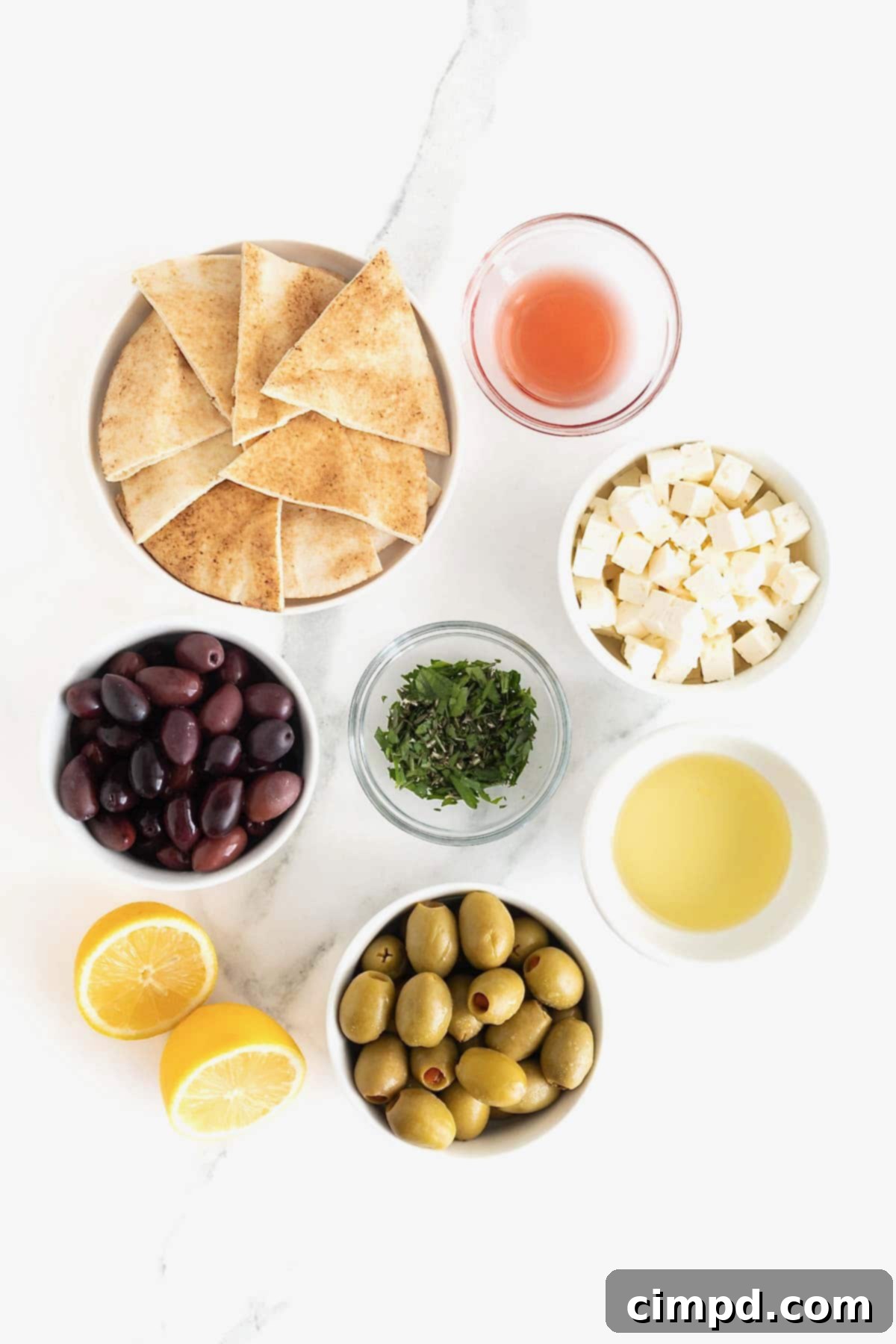 Ingredients for making marinated olives and feta laid out on a white surface, showing fresh herbs, olives, feta, lemon, and olive oil.