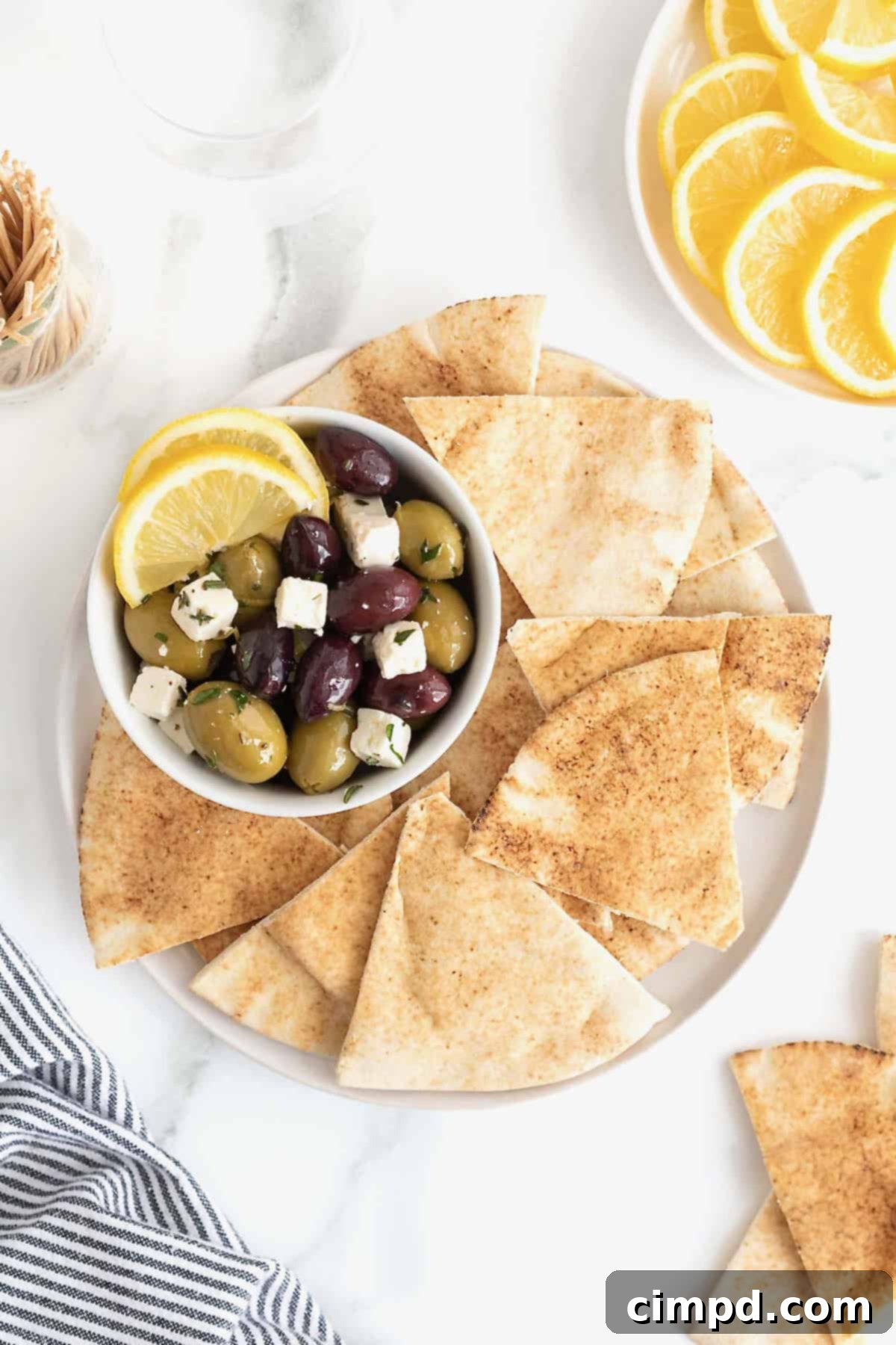 A bowl of marinated olives and feta surrounded by pita chips on a white serving platter, beautifully presented.