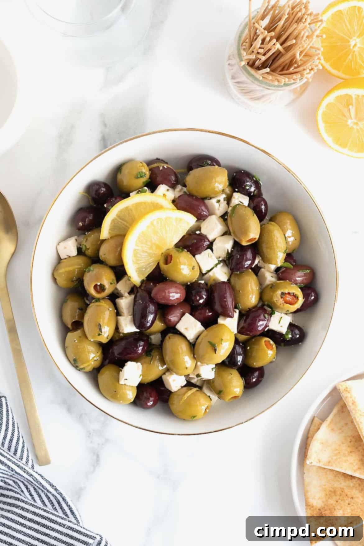 A gold rimmed white serving bowl filled with green and purple olives and small cubes of feta, a close-up highlighting the texture and freshness.