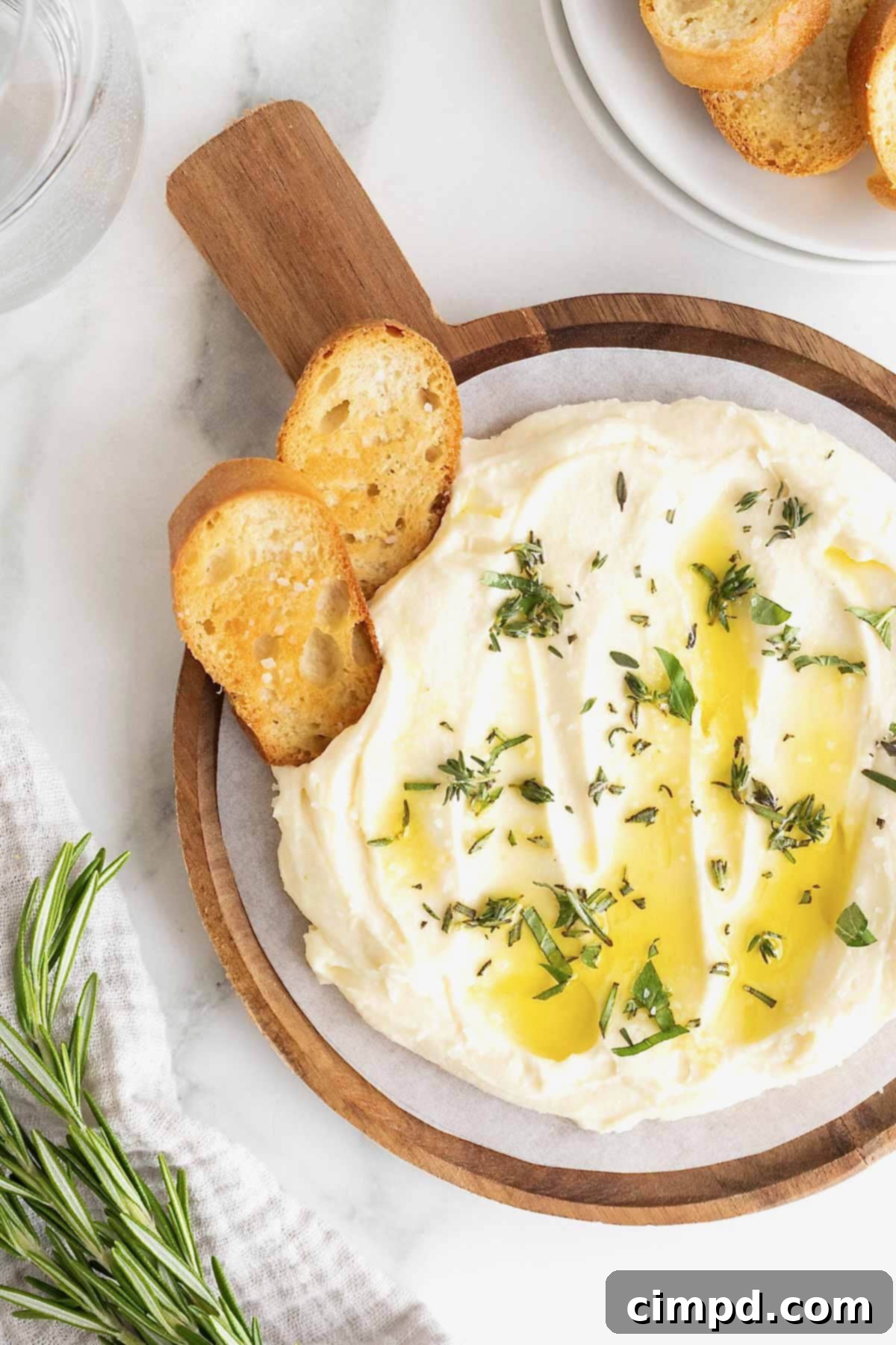 Creamy Whipped Brie 2 A beautiful wooden bowl filled with creamy whipped brie, generously drizzled with high-quality olive oil and garnished with fresh green herbs. Two pieces of crispy crostini are artfully placed within the dip, ready for serving.
