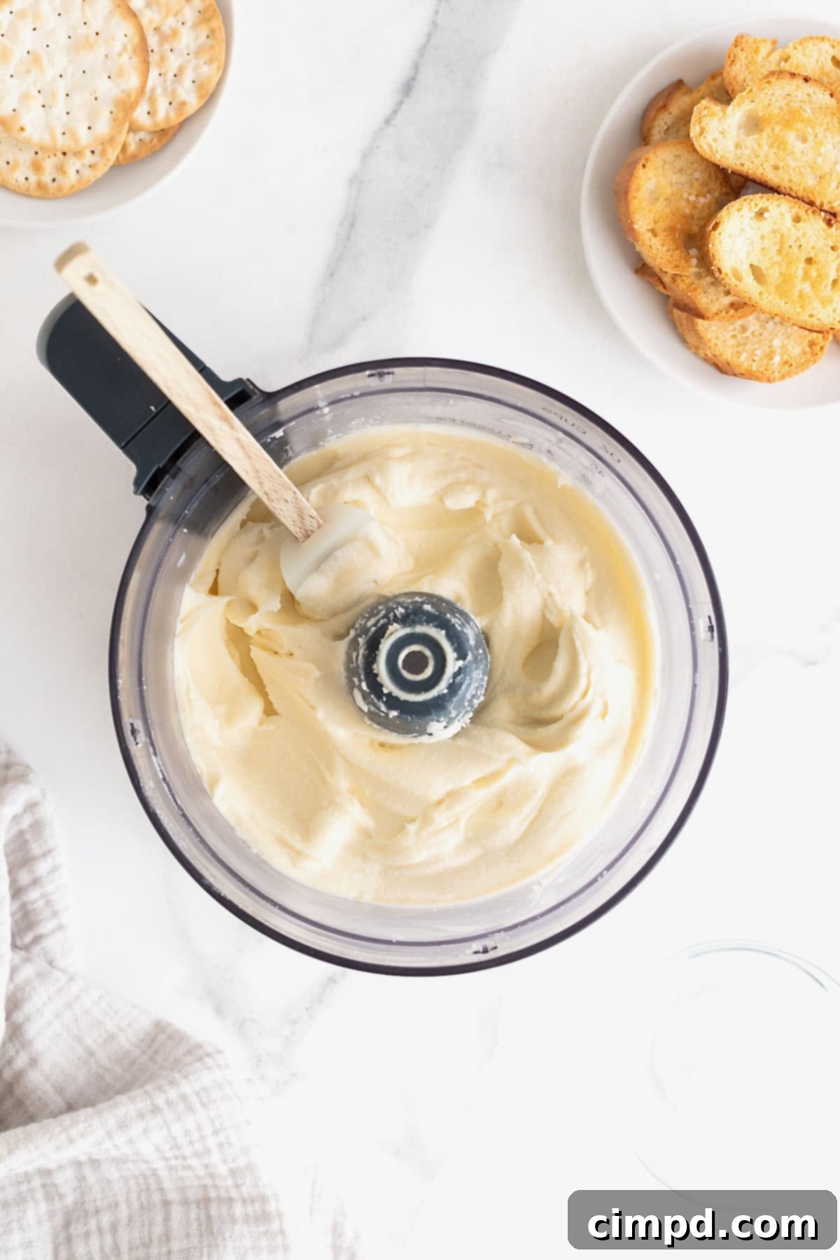 Creamy Whipped Brie 12 A food processor bowl filled with creamy, smooth whipped brie, accompanied by a wooden-handled spatula resting on the side, highlighting the velvety texture achieved during the blending process.