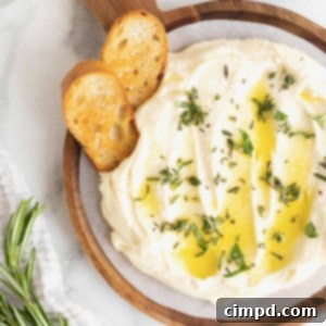 Creamy Whipped Brie 14 A wooden serving plate of fluffy whipped brie, elegantly drizzled with golden olive oil. Two pieces of crispy crostini are casually stuck in the brie, inviting immediate enjoyment.