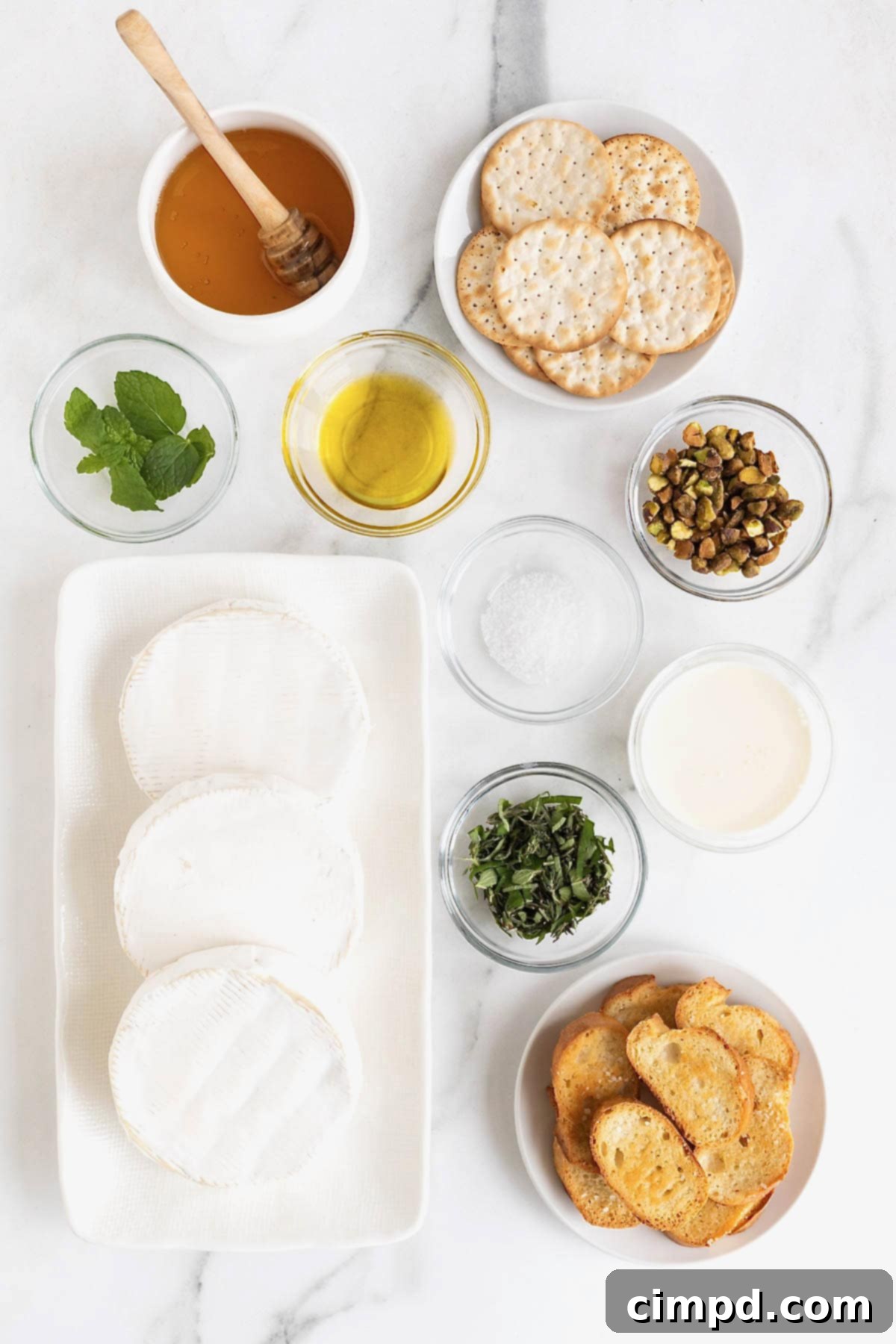 Creamy Whipped Brie 5 An inviting overhead view of small white dishes neatly arranged on a pristine white marble counter, each holding fresh ingredients essential for making whipped brie. Visible elements include creamy brie chunks, vibrant green herbs, golden olive oil, and a glistening drizzle of honey, all promising a delicious culinary creation.
