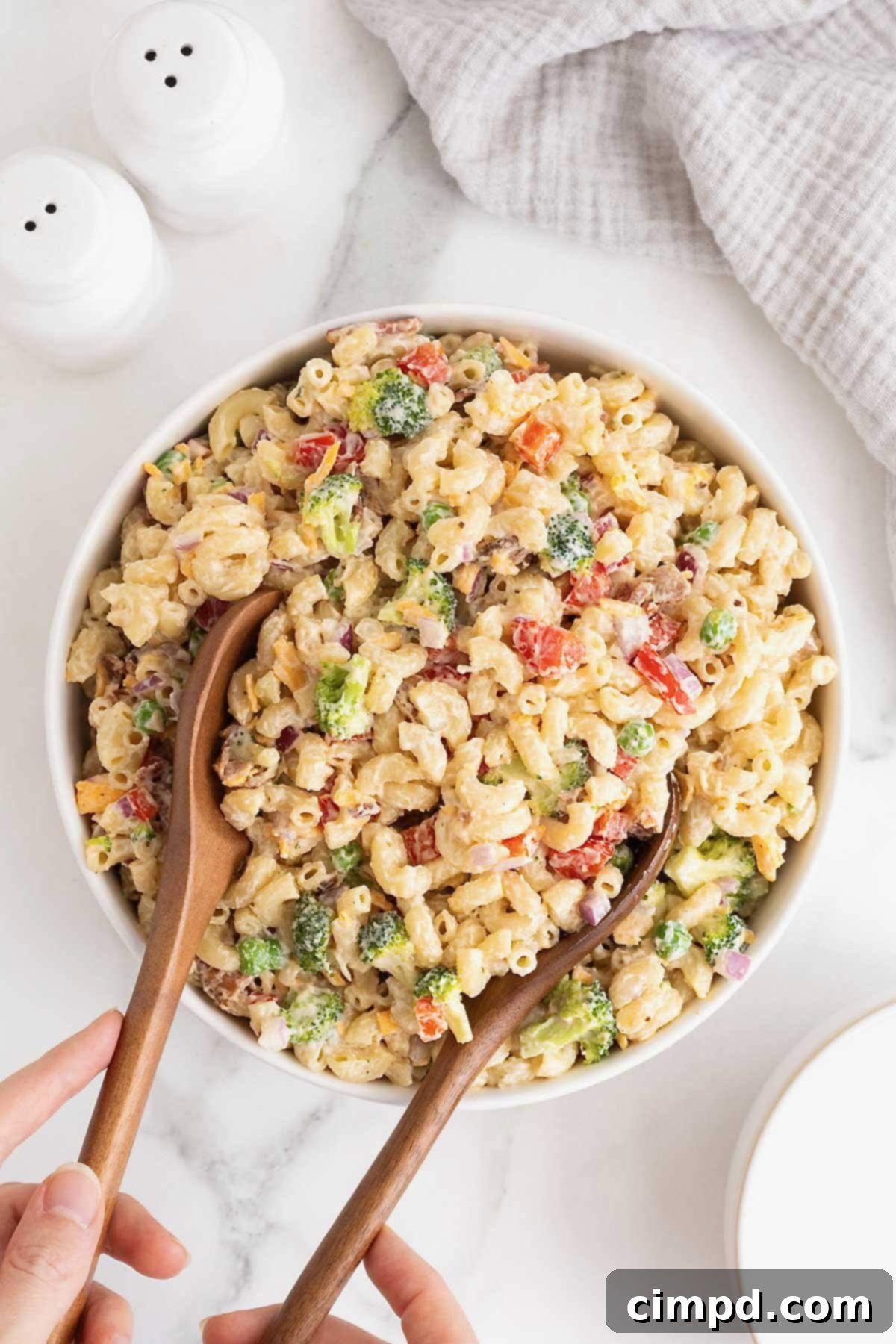 Two wooden serving spoons in a large white serving bowl of macaroni salad, highlighting fresh ingredients like sweet peas, red onion, and cheese.
