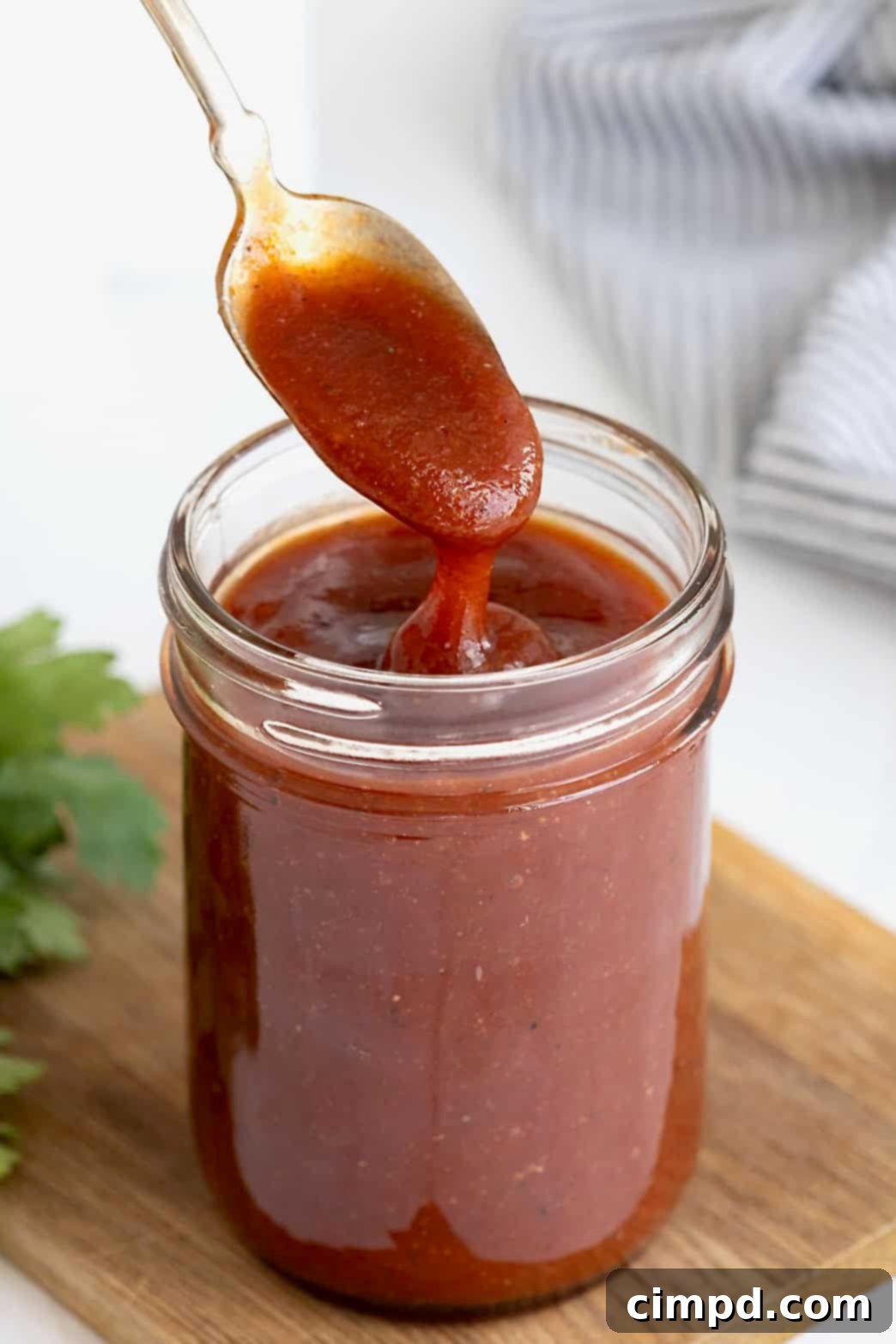 Handcrafted Barbecue Sauce 2 A glistening spoonful of rich, homemade barbecue sauce being lifted from a wide-mouth mason jar, resting on a rustic wooden cutting board.