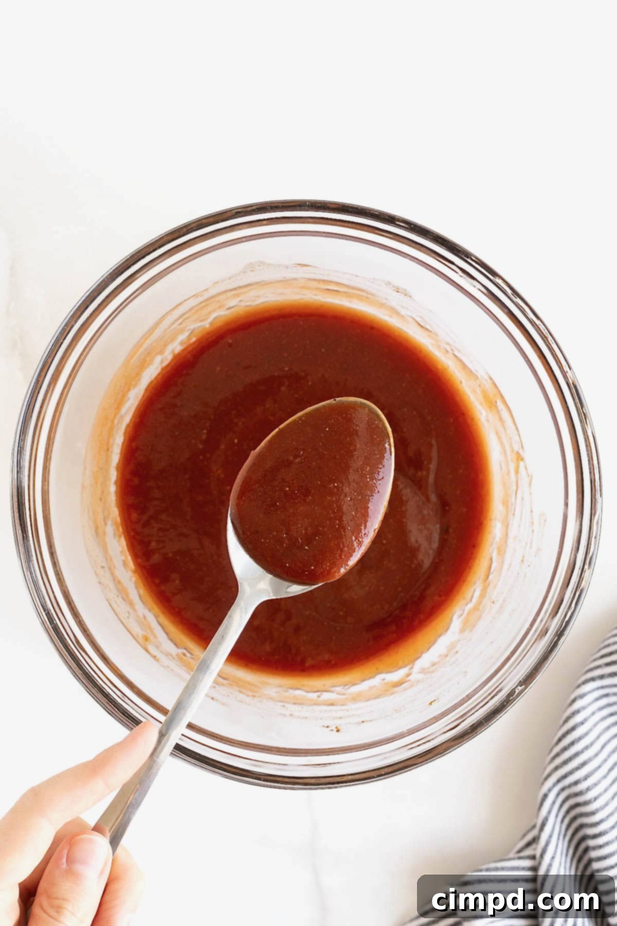 Handcrafted Barbecue Sauce 4 A silver spoon dipping into a large, clear glass mixing bowl filled with freshly made, smooth homemade barbecue sauce.