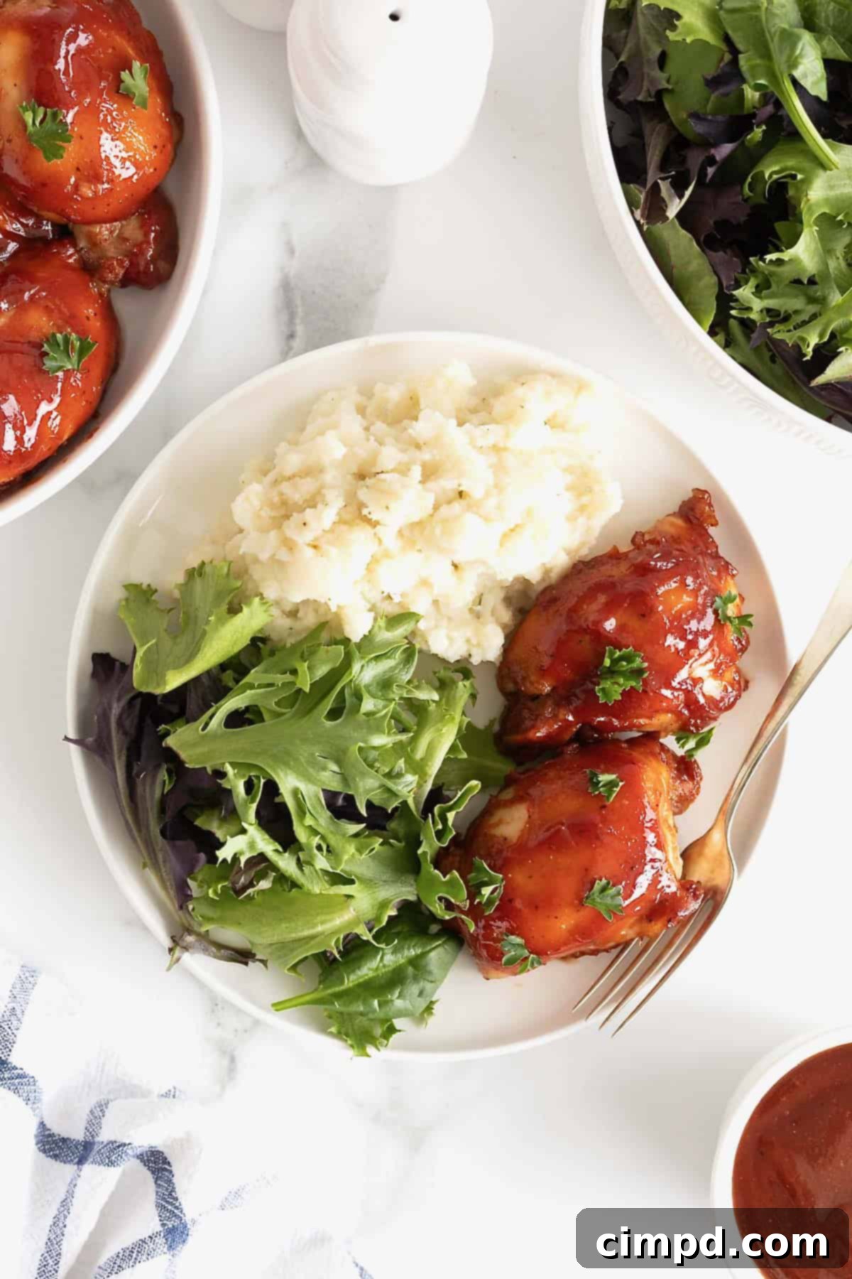 A white dinner plate with two bbq chicken thighs, a green salad, and mashed potatoes.
