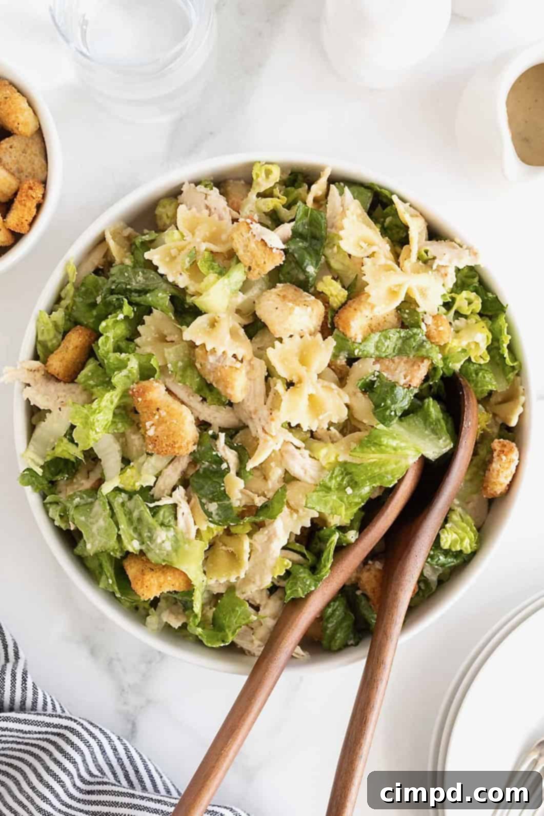A large white salad bowl of caesar pasta salad with chicken. There are two wooden serving spoons in the bowl. 