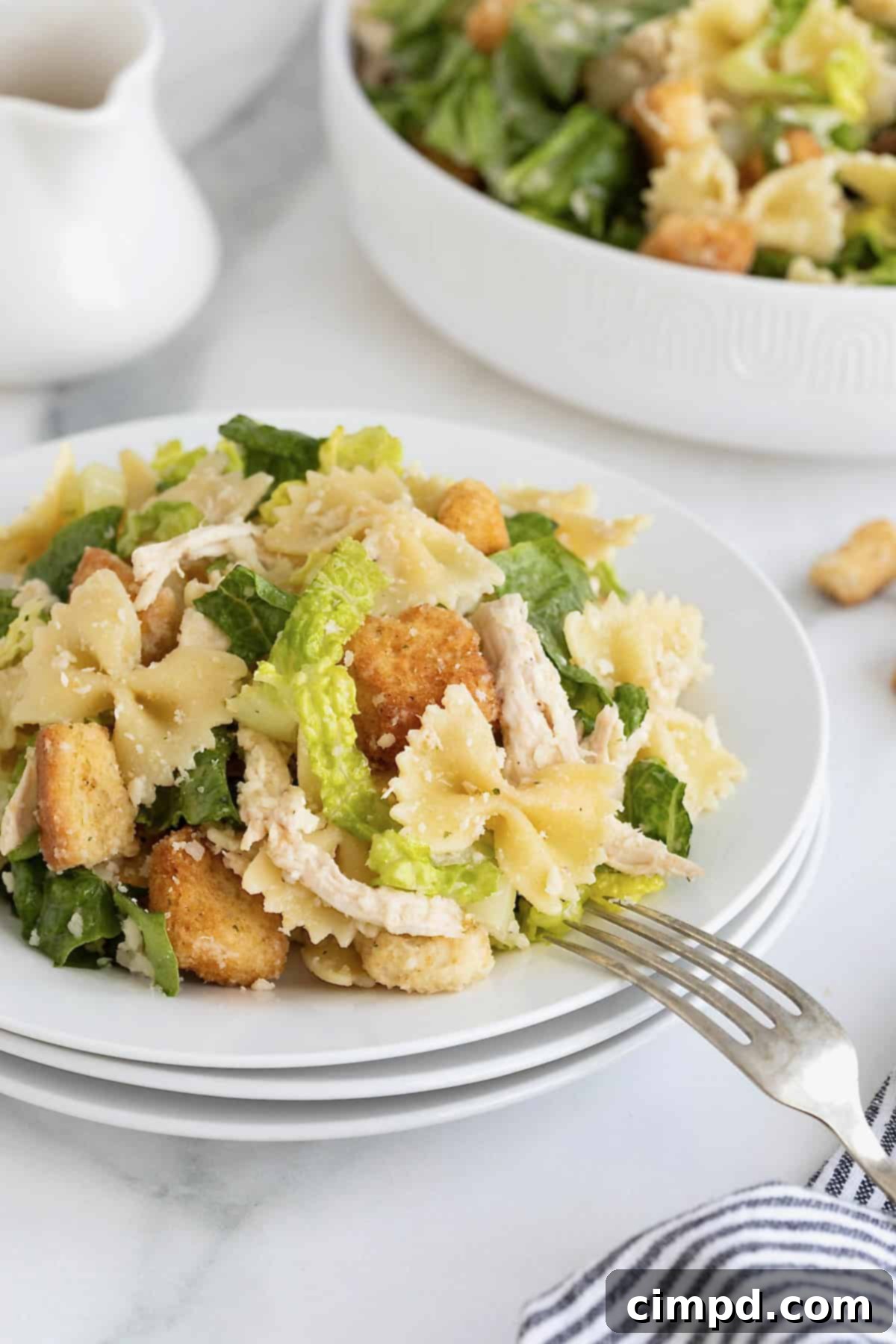 A stack of three white salad plates with a serving of chicken caesar pasta salad on the top plate. A fork is resting on the plate.