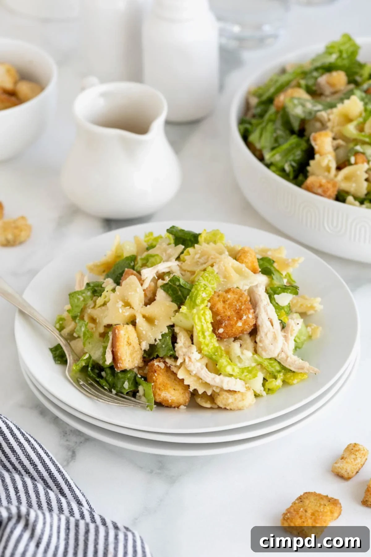 A stack of three white salad plates with a serving of chicken caesar pasta salad on the top plate. A fork is resting on the plate.