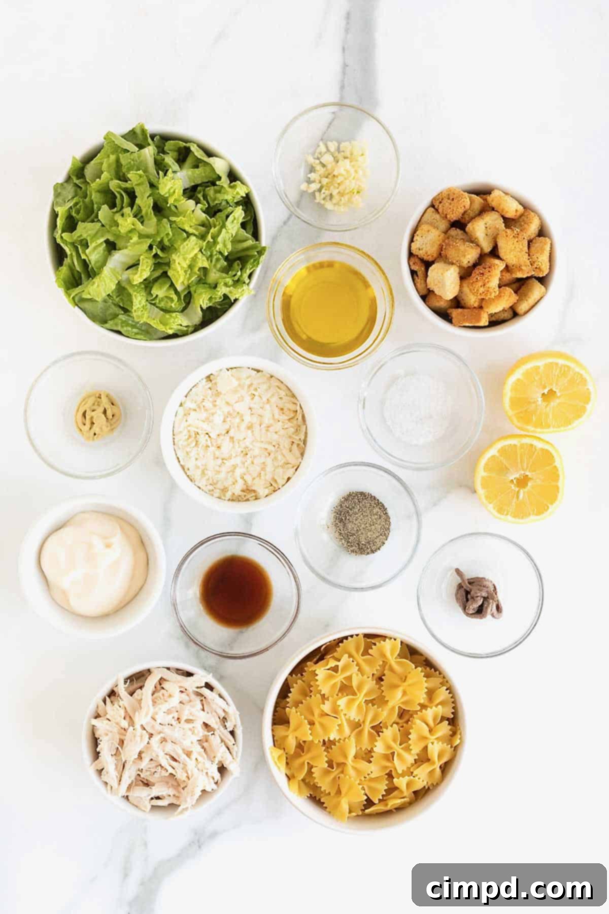 Ingredients to make chicken Caesar pasta salad in small glass dishes on a white marble counter.