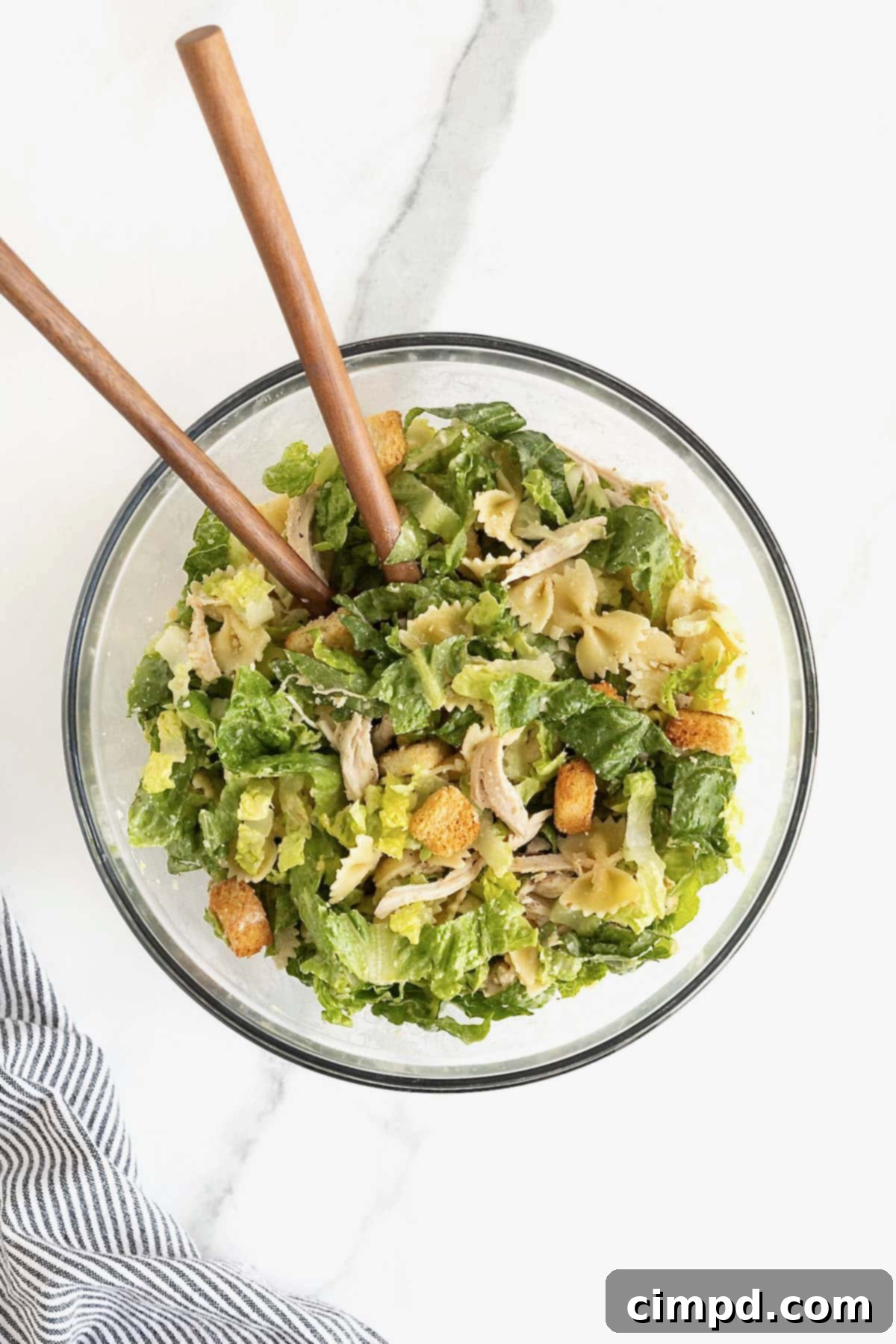 A large glass mixing bowl of caesar pasta salad with chicken. There are two wooden serving spoons in the bowl.