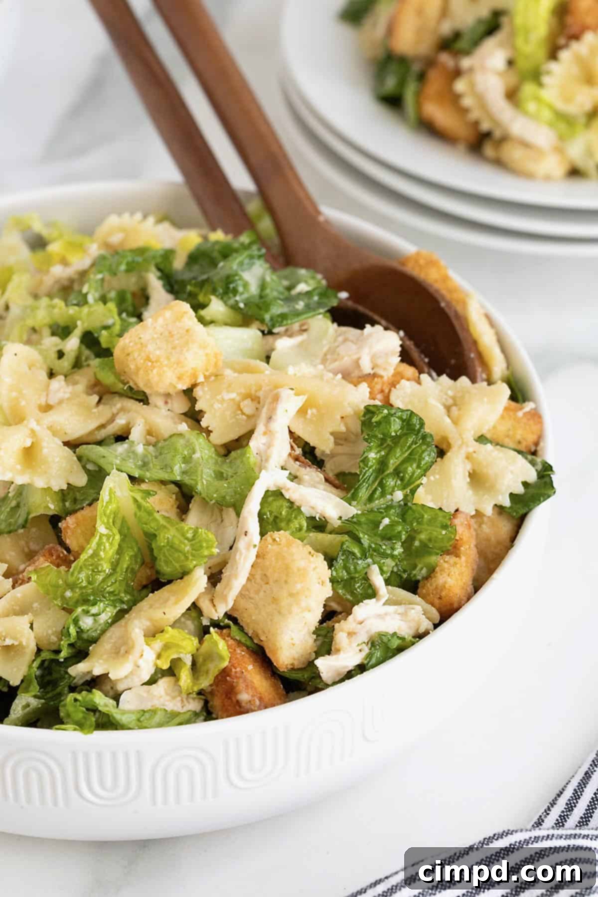 A large white salad bowl of caesar pasta salad with chicken. There are two wooden serving spoons in the bowl.