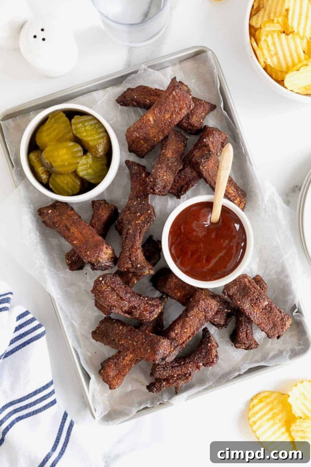 A parchment lined aluminum baking sheet of baby back ribs, ready to be served. There is a small white dish of pickles at the top left and a small white dish of barbecue sauce in the center, inviting dipping.
