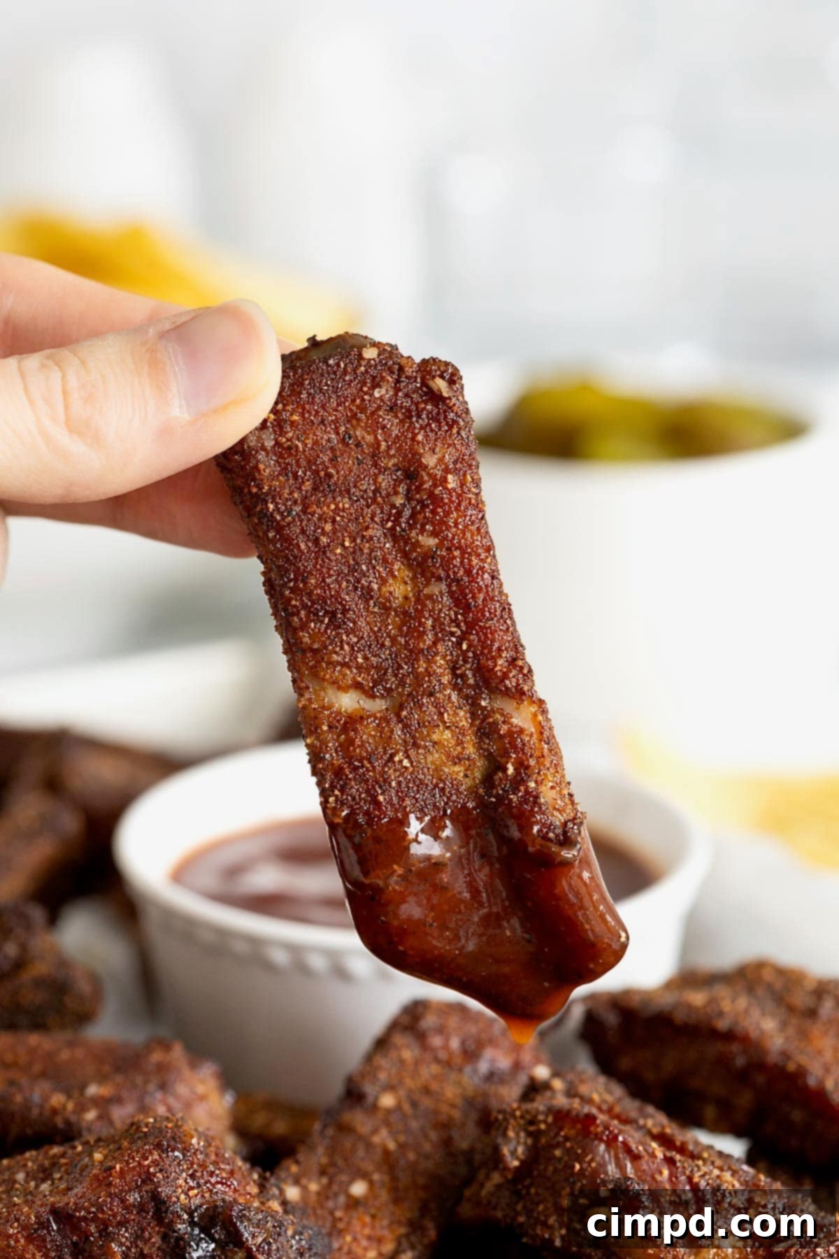 A succulent baby back rib being dipped into a small white bowl of rich barbecue sauce, showcasing its inviting color and texture.