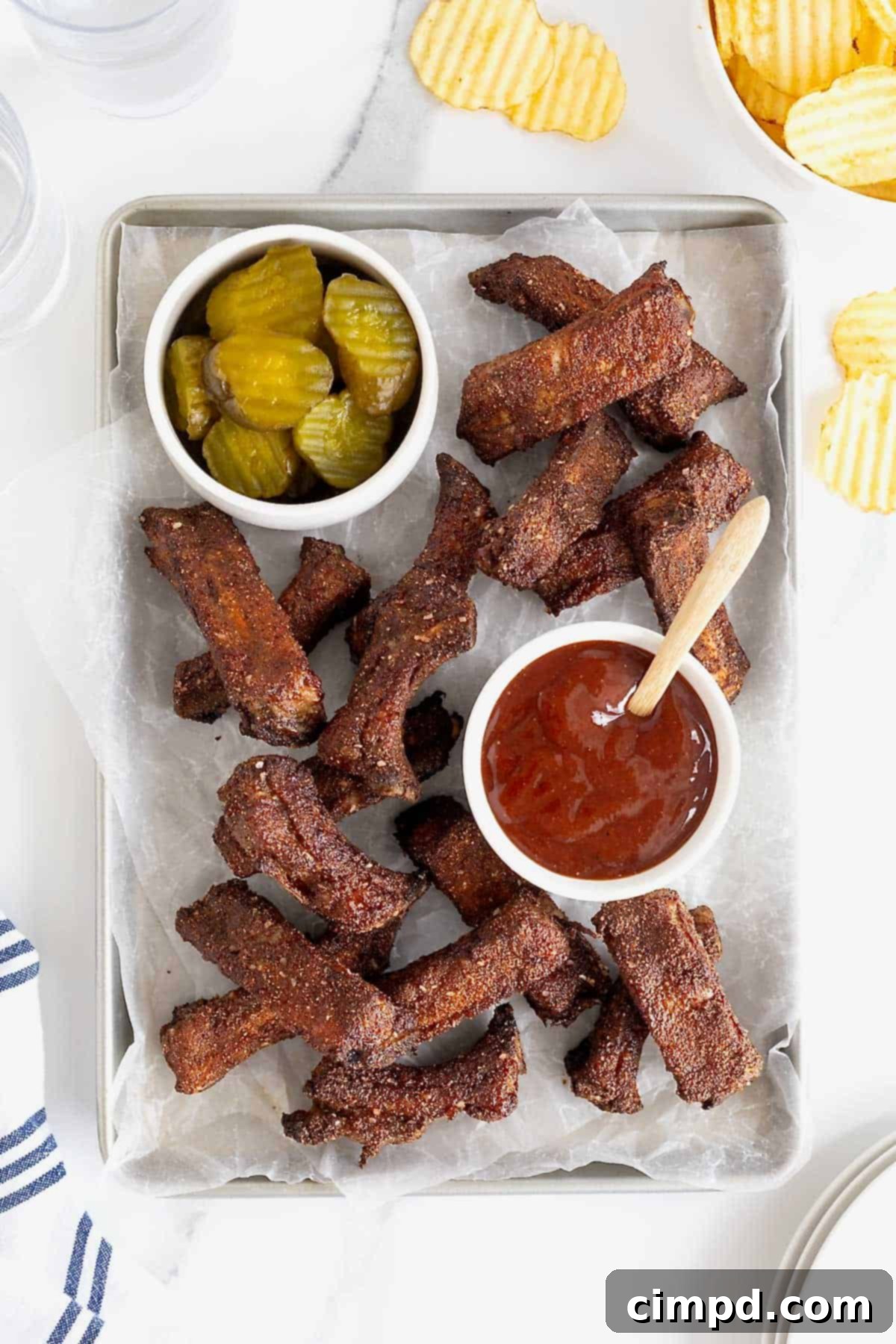 A full parchment lined aluminum baking sheet filled with golden-brown, dry-rubbed baked baby back ribs, accompanied by small dishes of pickles and barbecue sauce.