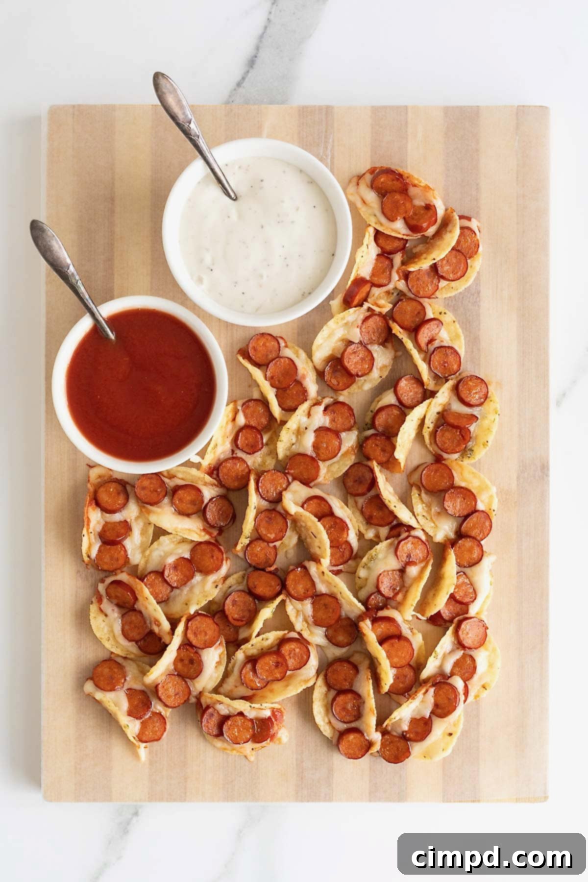A pile of pizza taco bites on a parchment lined wooden cutting board. There is a small white bowl of pizza sauce and a small white bowl of ranch dressing on the top left corner of the board.