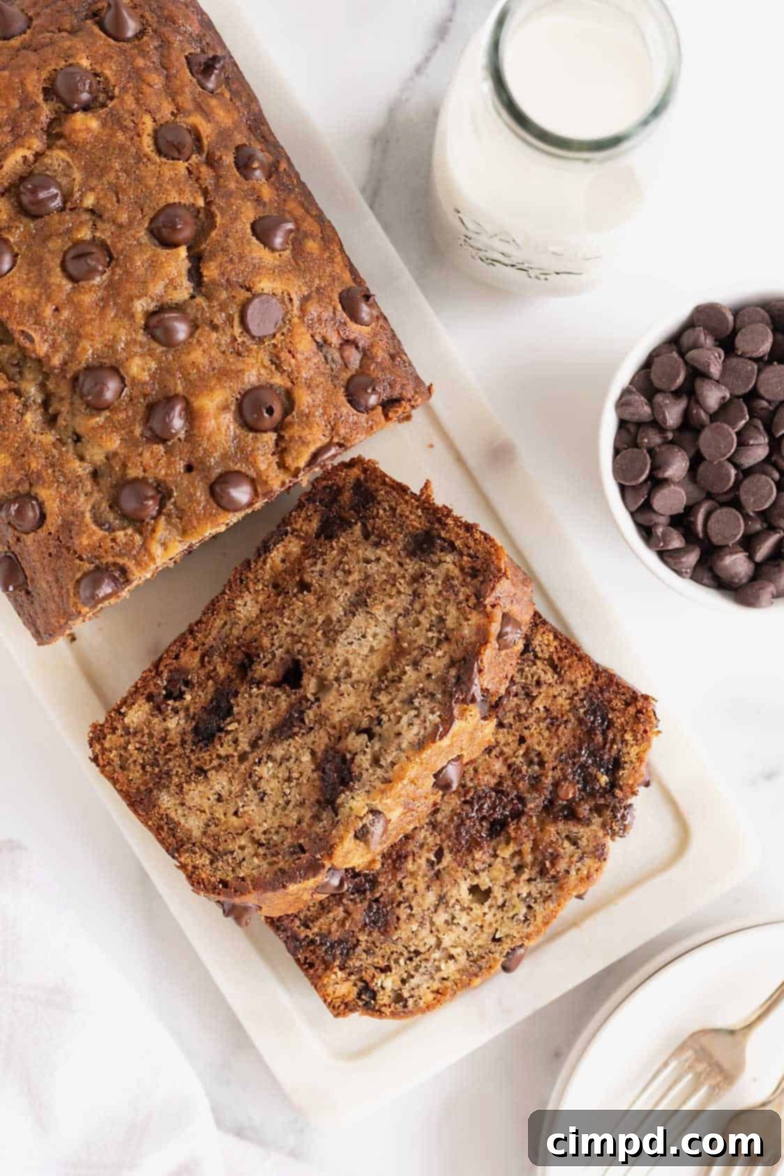 A loaf of chocolate chip banana bread on a marble cutting board. A small white dish of chocolate chips sits to the right side.