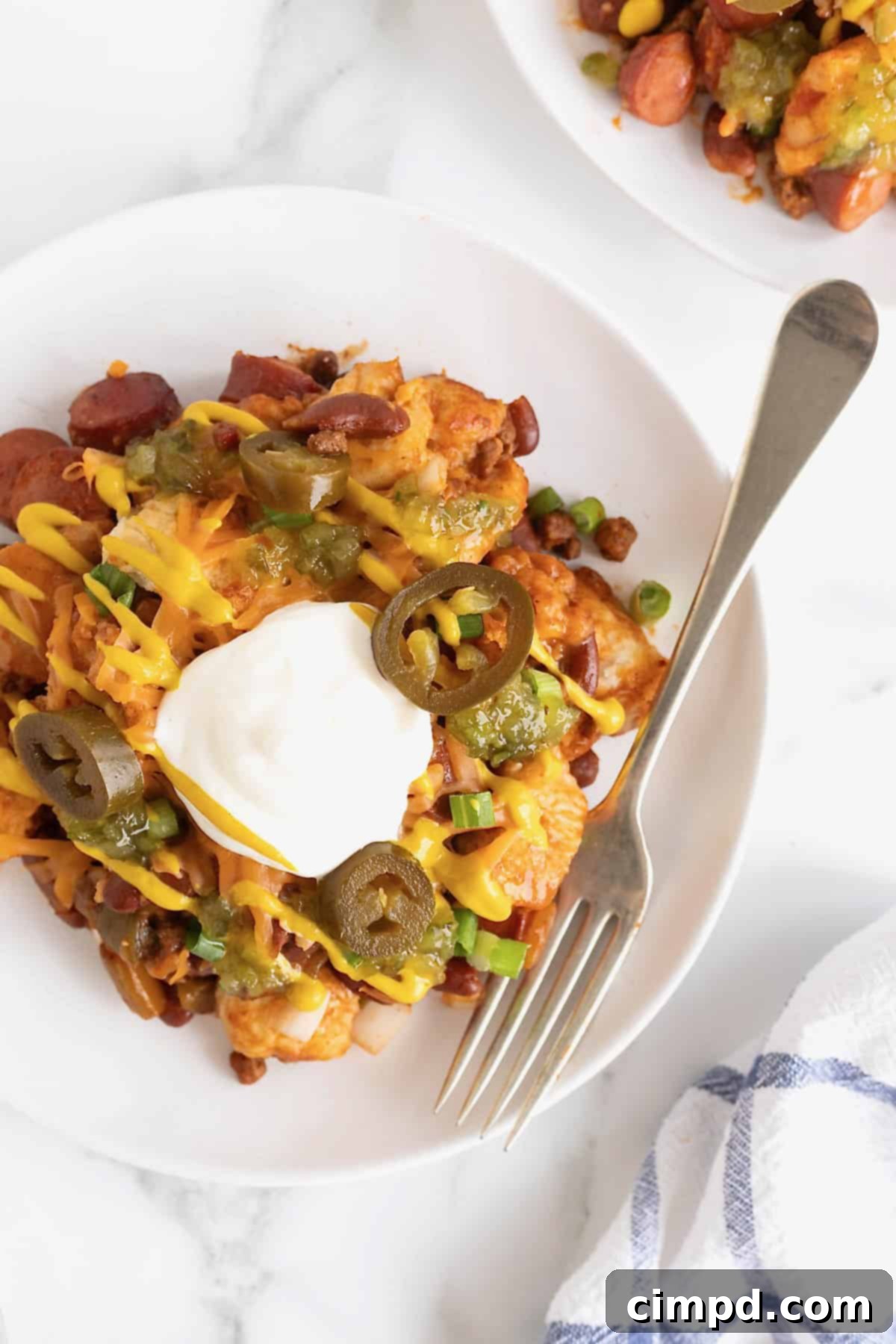 A serving of chili cheese dog casserole on a white plate, topped with a drizzle of mustard, a dollop of sour cream, and four jalapeño slices.
