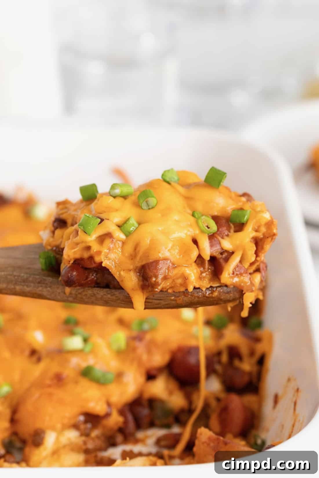 A wooden spoon lifting a serving of chili cheese dog casserole from a white casserole dish, showing the cheesy, chili-covered hot dogs and bun pieces.