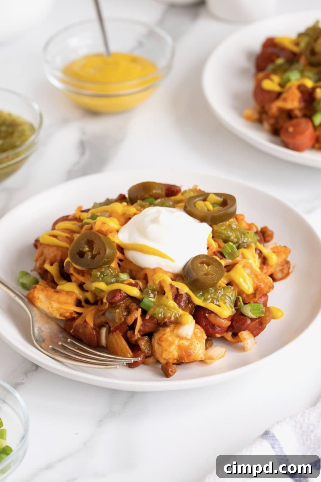 A serving of chili cheese dog casserole topped with a drizzle of mustard, a dollop of sour cream, and four jalapeño slices on a white plate, ready to be enjoyed.