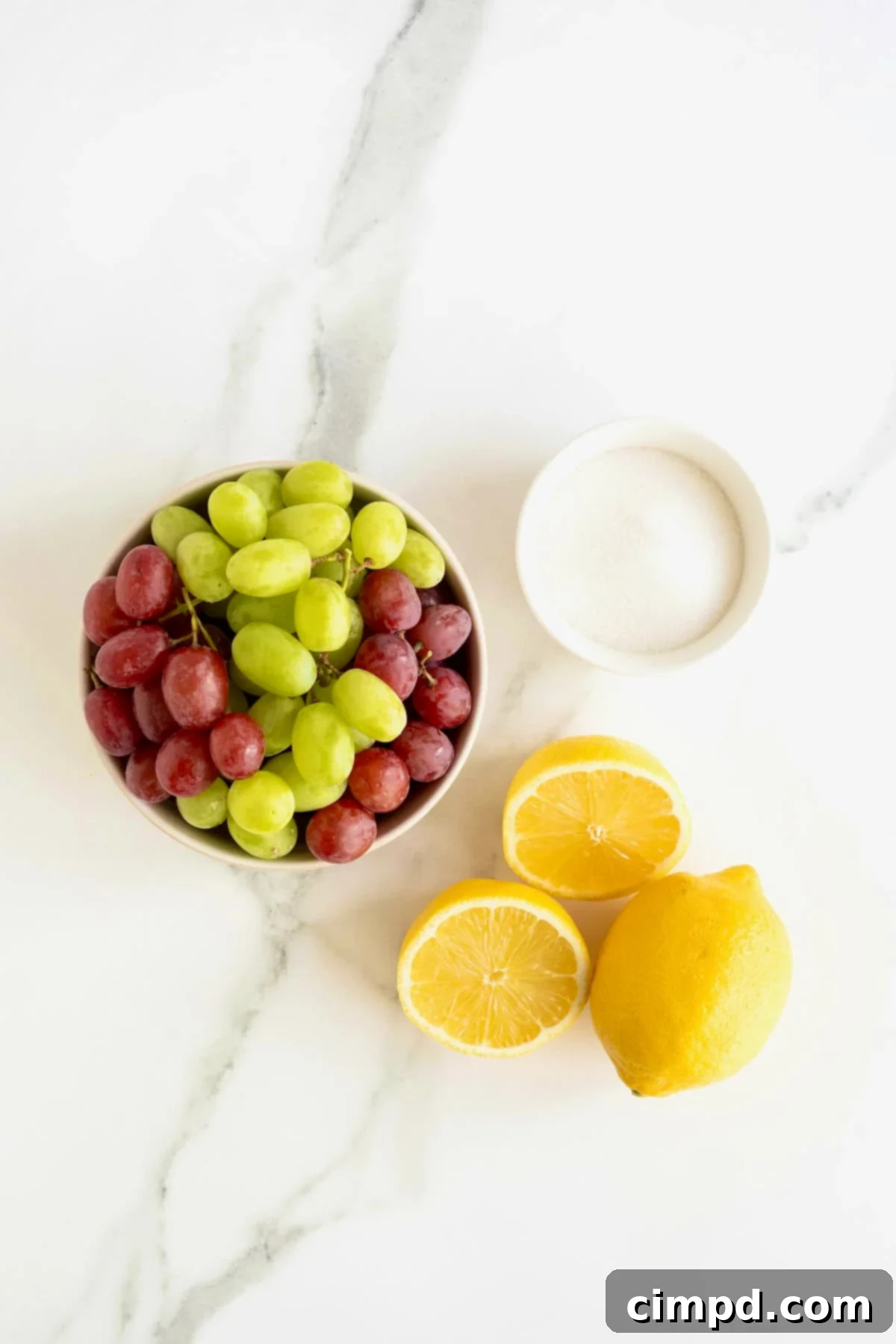 Effortless Homemade Grape Jam 4 A collection of fresh green and red seedless grapes, granulated sugar, and fresh lemons laid out on a white marble counter, showcasing the simple ingredients for homemade grape jam.