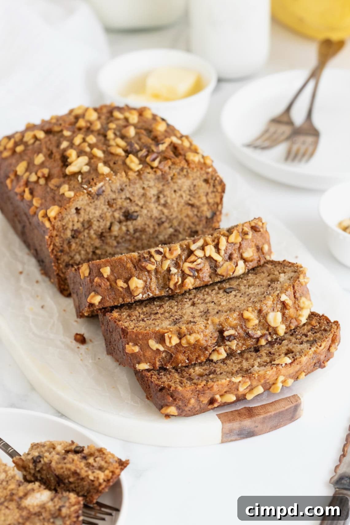 A golden-brown loaf of homemade brown butter banana nut bread on a rustic white cutting board. Three thick slices are cut, revealing a moist, dense crumb studded with walnuts.