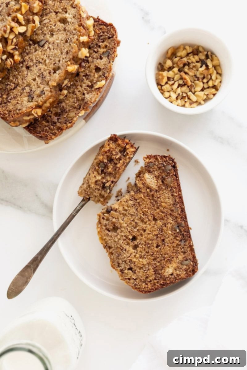 A delicious slice of brown butter banana nut bread on a white rimmed plate, with a fork poised next to it, inviting a bite.
