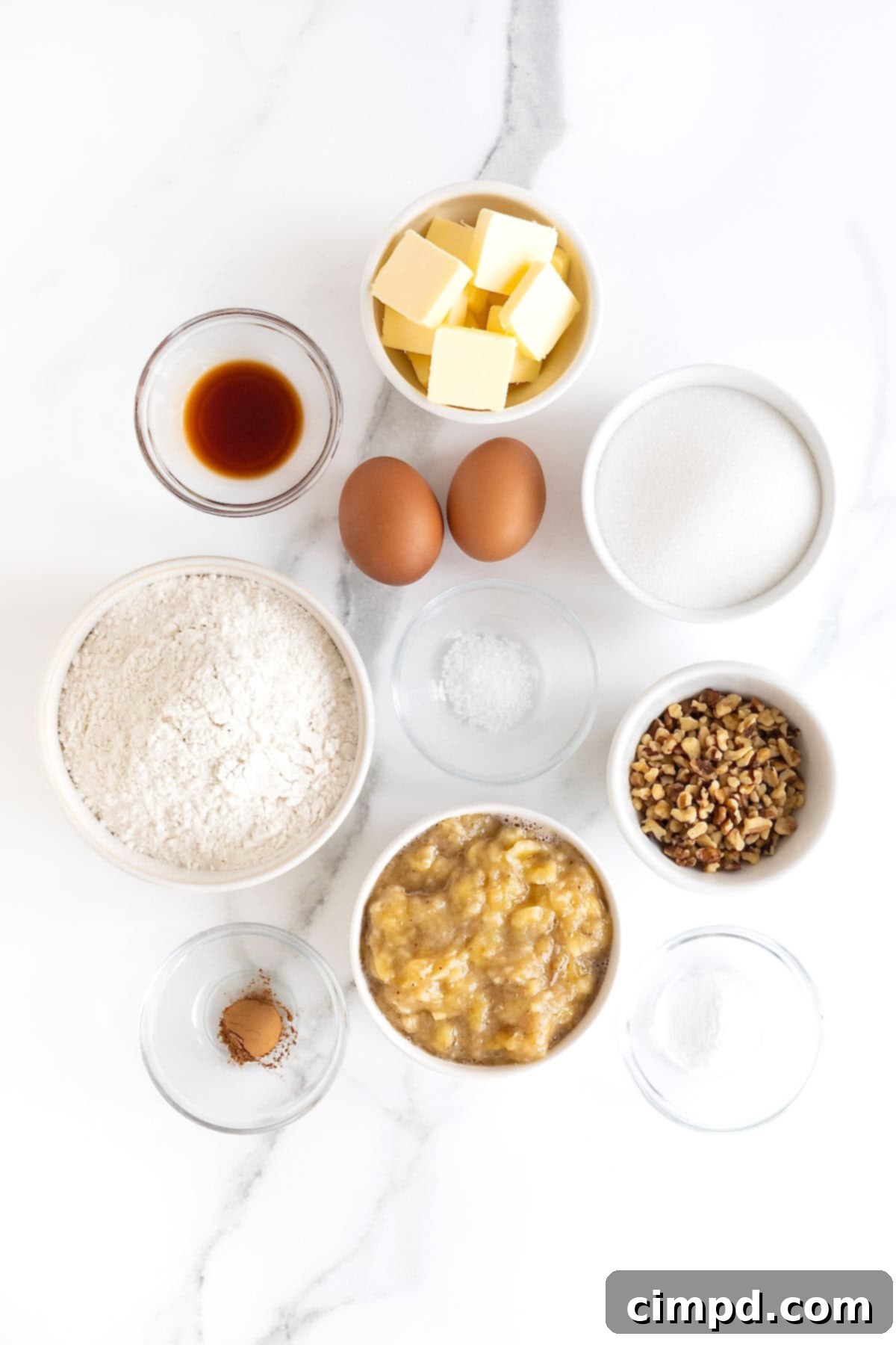 A selection of fresh ingredients neatly arranged in small white dishes on a white marble counter, ready to be used for making brown butter banana nut bread.