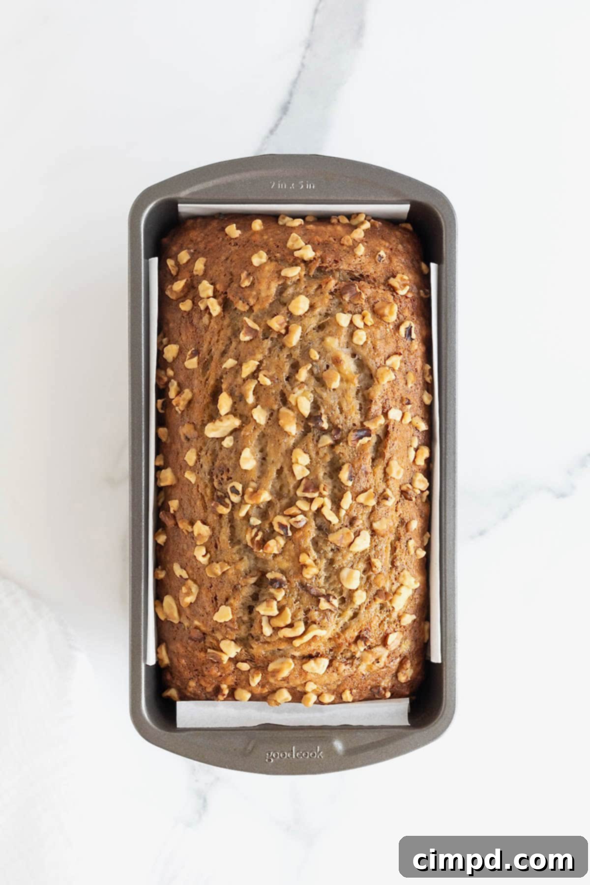 A perfectly baked loaf of brown butter banana nut bread resting in a dark aluminum loaf pan on a cool white marble countertop, having just come out of the oven.