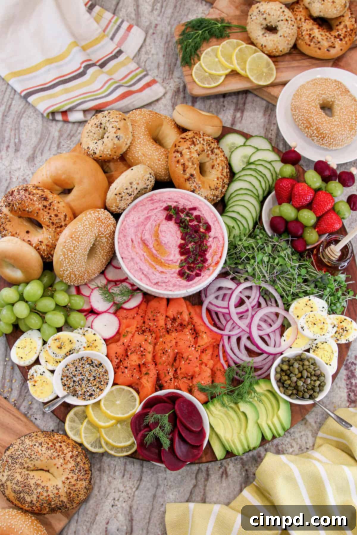 A large round wooden board artfully arranged with various bagel sandwich fixings, including bagels, vegetables, lox, and a central white bowl of bright pink beet cream cheese spread.