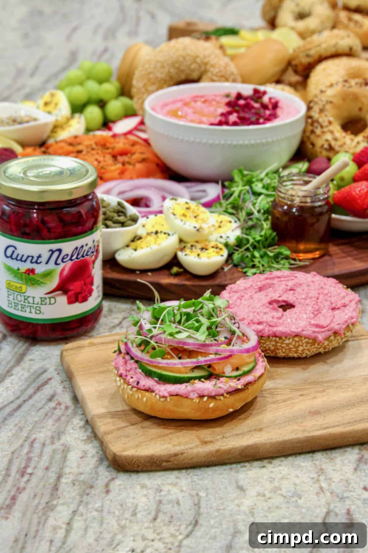 A close-up of a beautifully assembled bagel breakfast sandwich on a small wooden board. It features a generous smear of pink beet cream cheese spread, fresh vegetables, and smoked salmon, with a jar of pickled beets in the background.