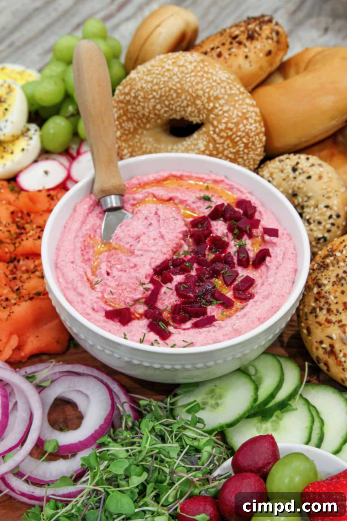 A white bowl of bright pink beet cream cheese spread, garnished with fresh dill and honey, sits in the center of a breakfast bagel sandwich board. The board is laden with a variety of colorful toppings, ready for assembly.
